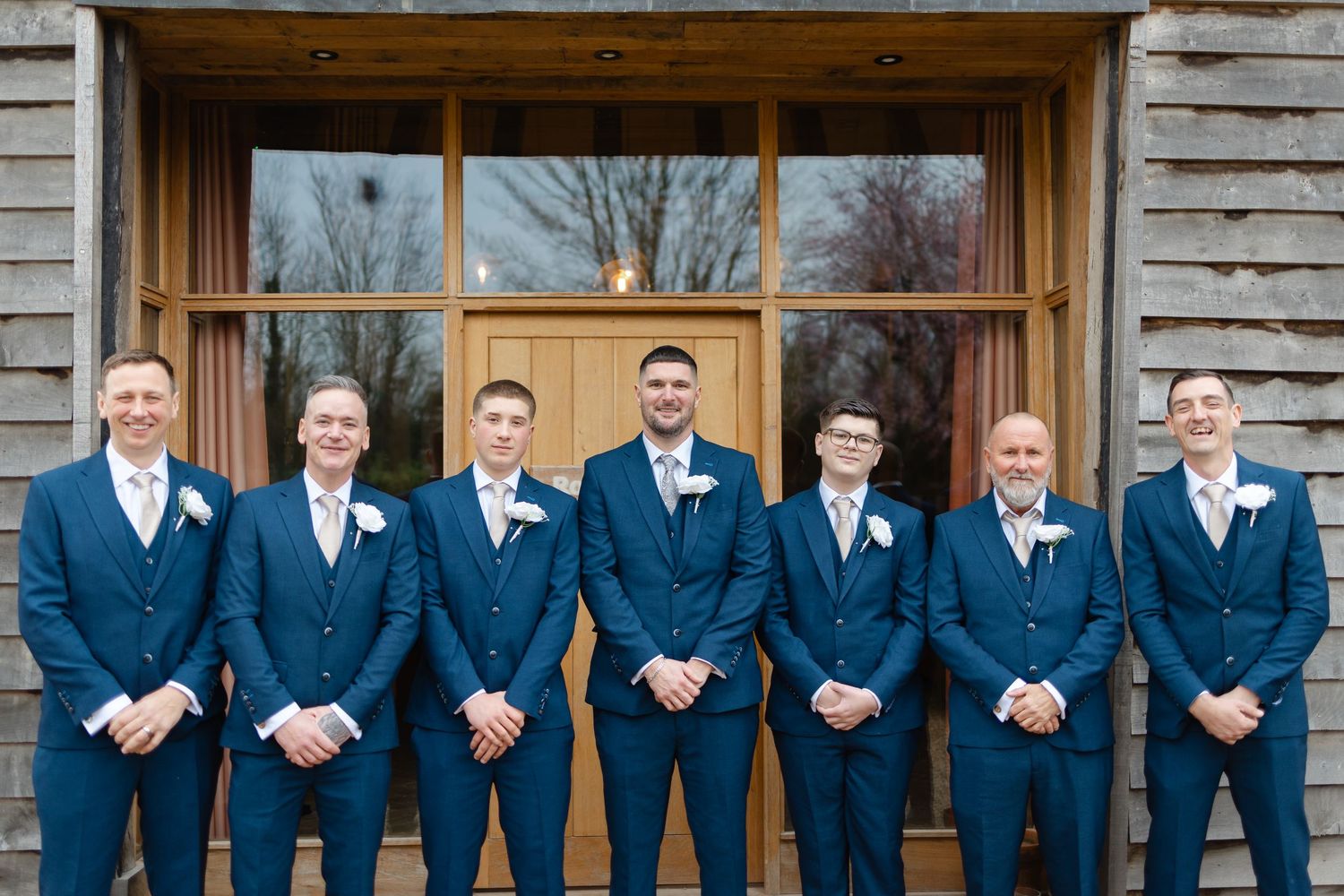 Wedding party members wearing navy blue suits pose in front of rustic building.