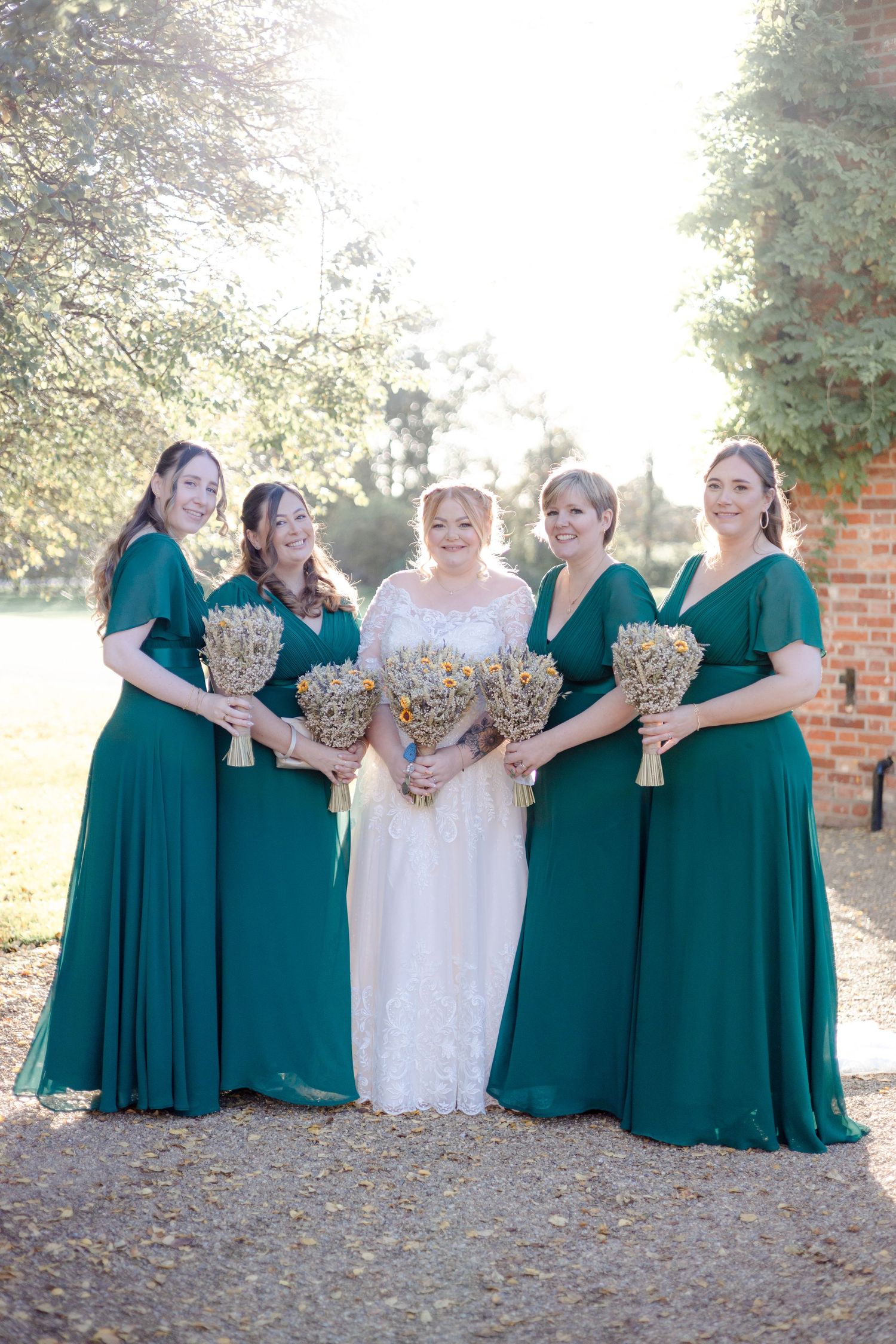 Bridesmaids in emerald green dresses pose with bride in white gown during outdoor wedding photoshoot.