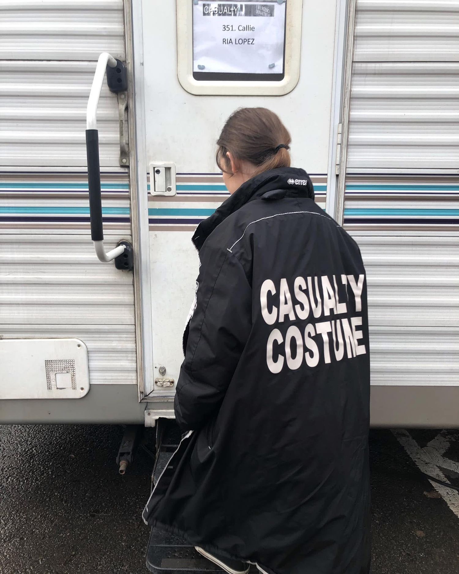 A person in a black jacket with 'CASUAL COSTUME' text on the back is shown from behind near a trailer.