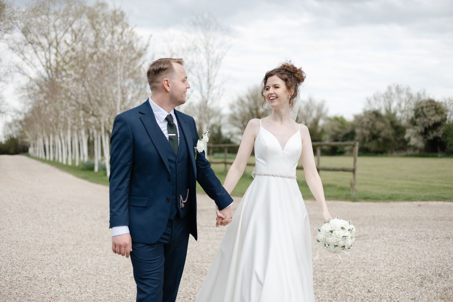 Candid wedding photos showing playful moments between couple on gravel path surrounded by white trees.