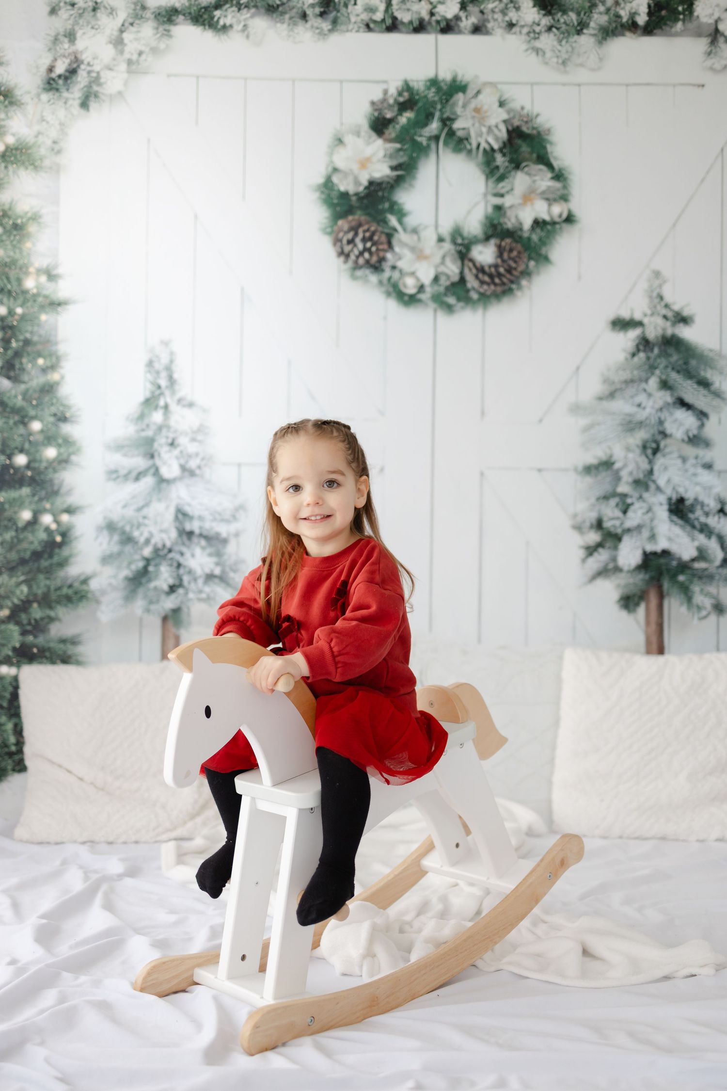 A child in a red dress rides a white rocking horse against a winter-themed backdrop.