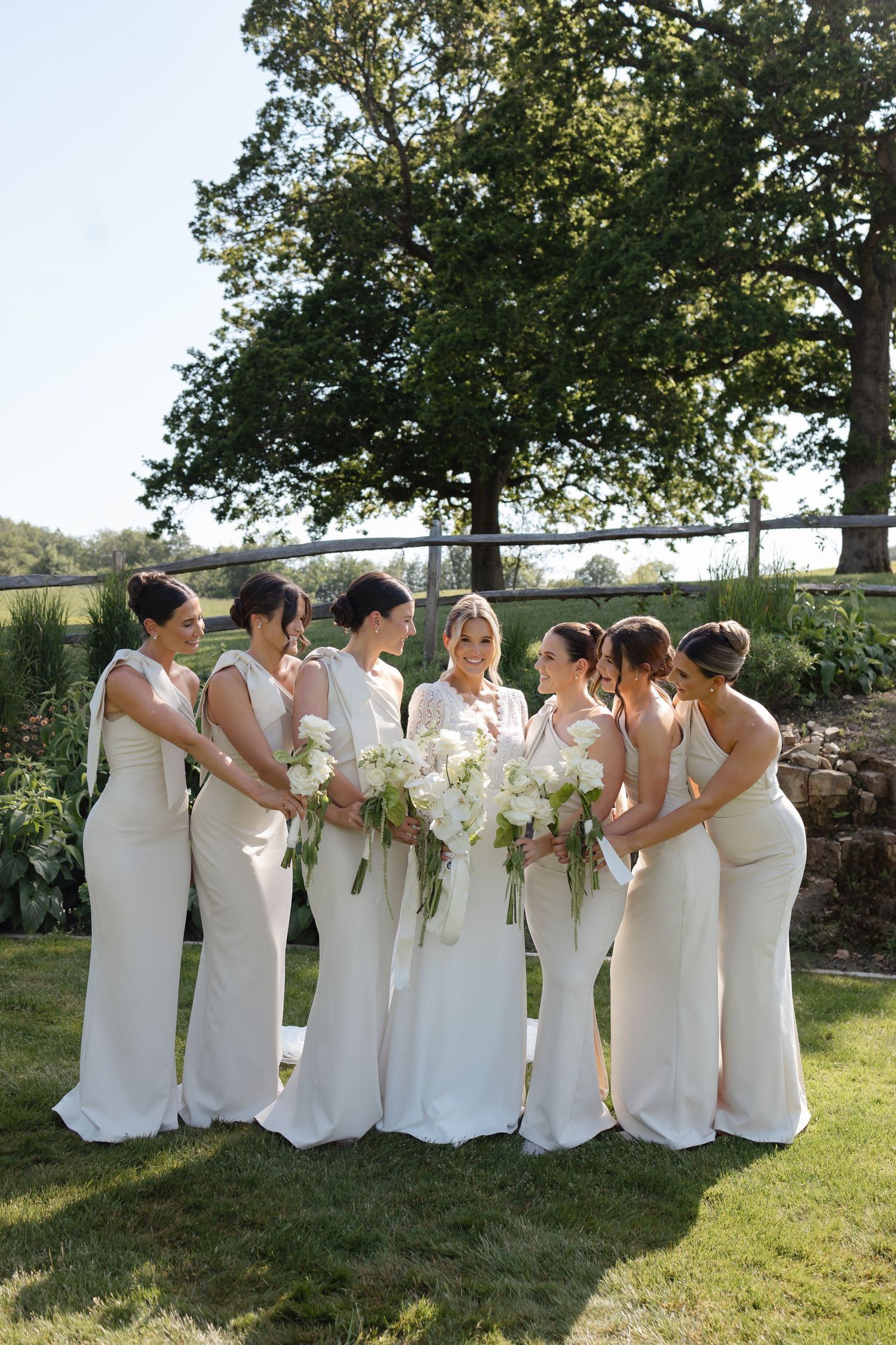 Bride with six bridesmaids in ivory gowns holding white floral bouquets outdoors on a sunny wedding day.