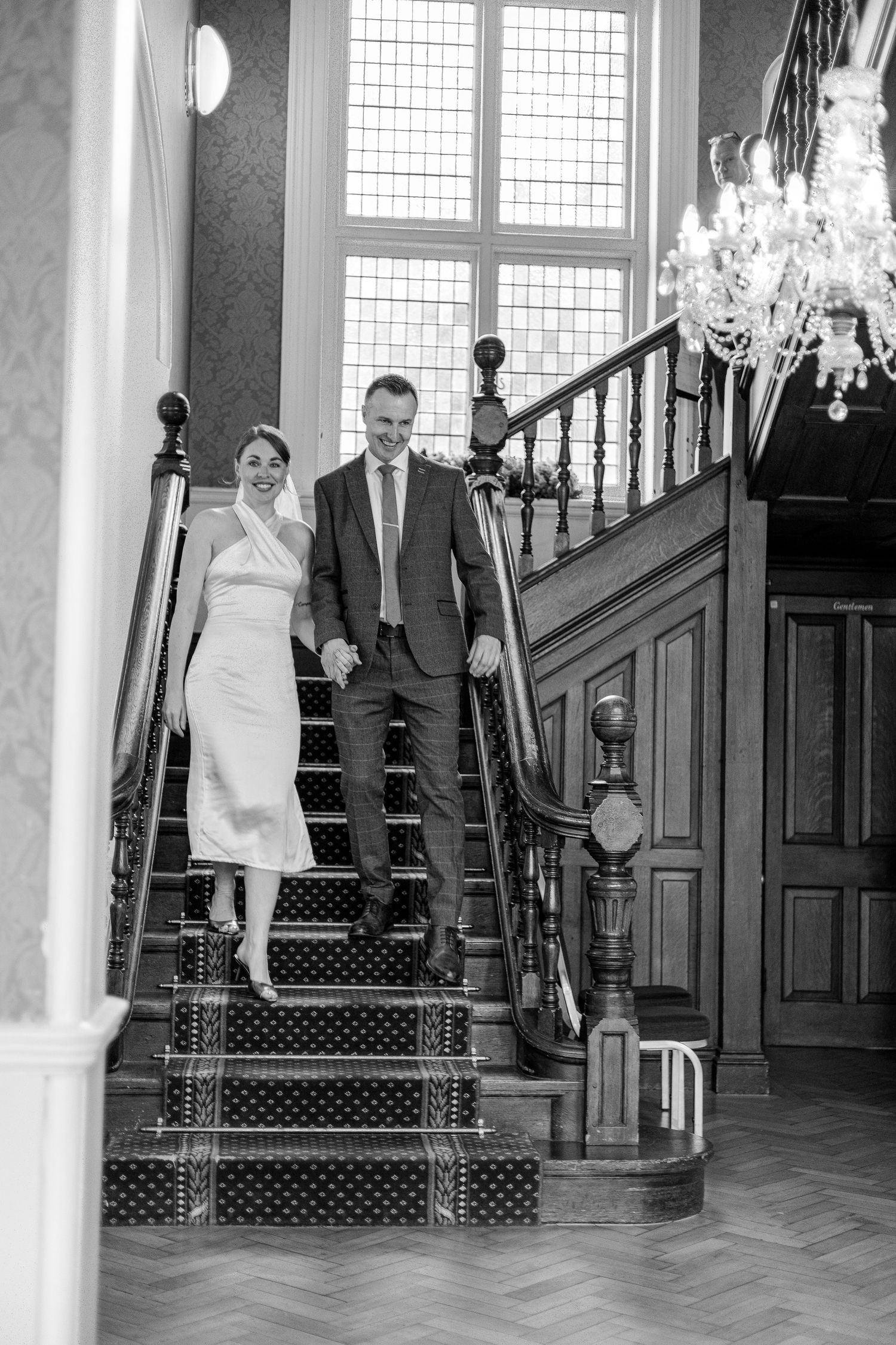 Couple descending elegant staircase with chandelier in grand indoor setting.