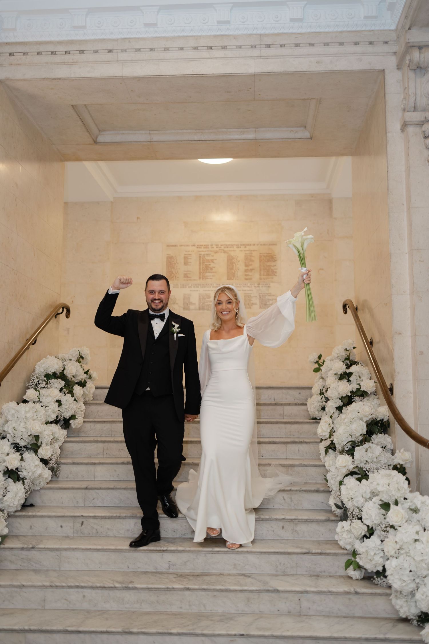 Wedding couple posing playfully on grand marble stairs decorated with white floral arrangements.