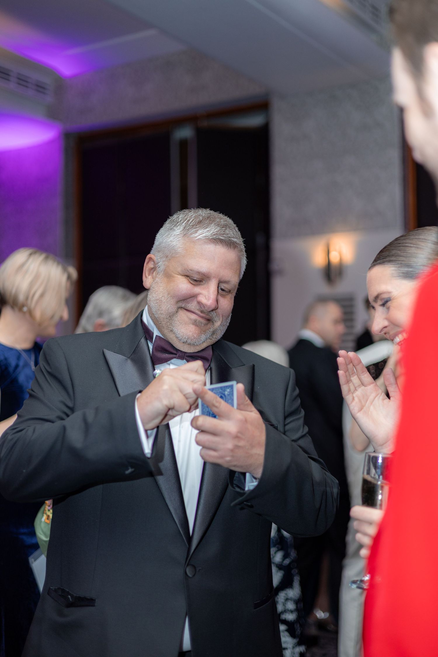 A magician in a black suit performs card tricks at an elegant evening event with purple lighting.
