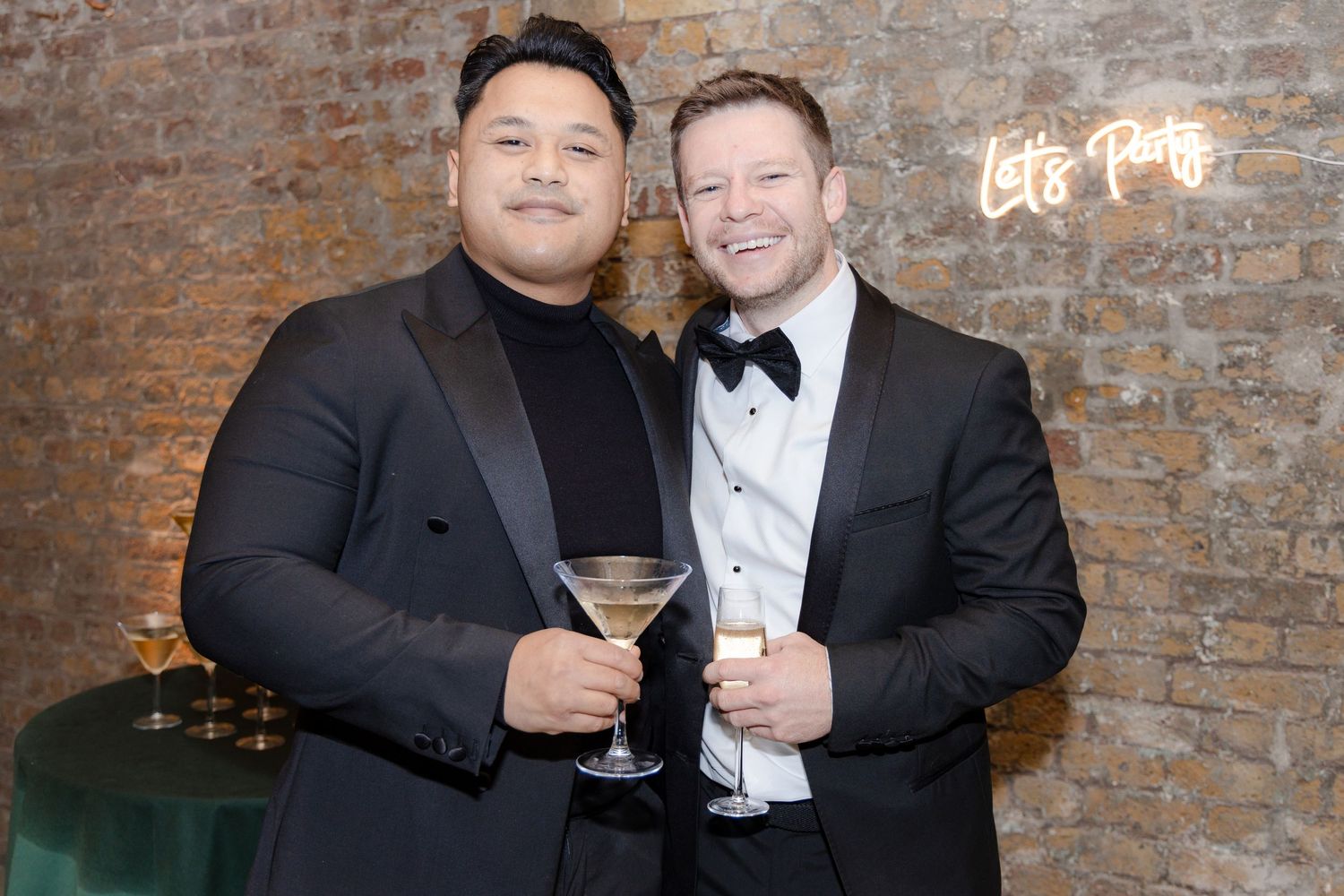 Two guests in formal attire holding martini glasses at an event with brick wall backdrop.