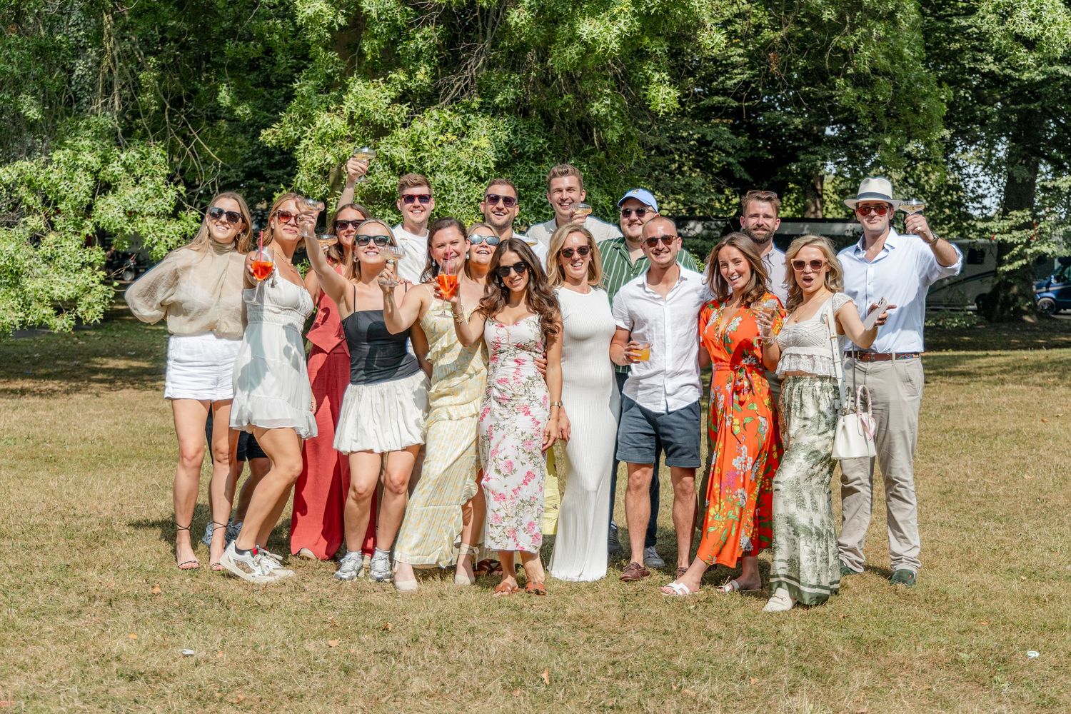 Large group of wedding guests in summer attire posing outdoors on grassy lawn.