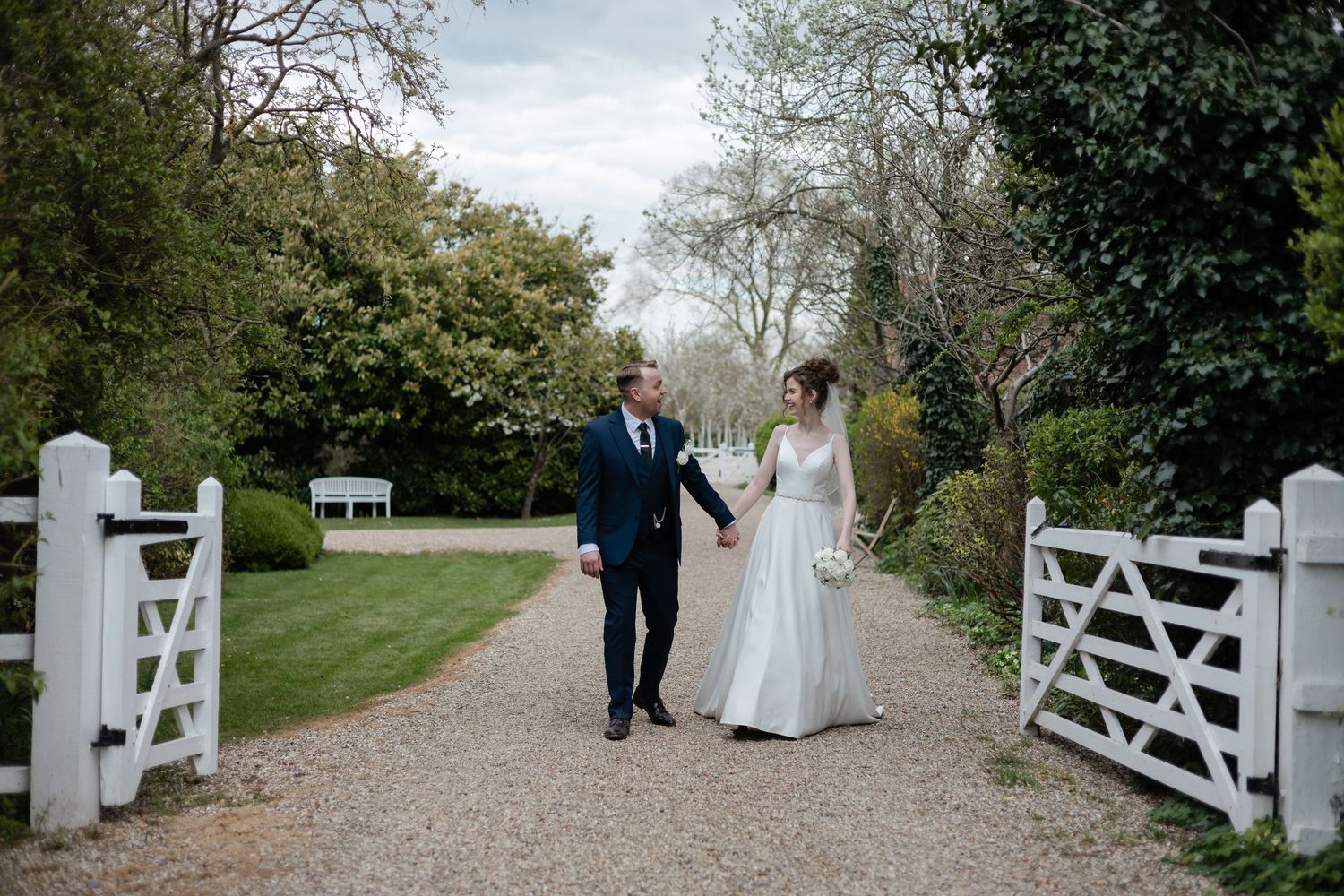 Romantic wedding couple walking and talking beside decorative white fence on garden path.