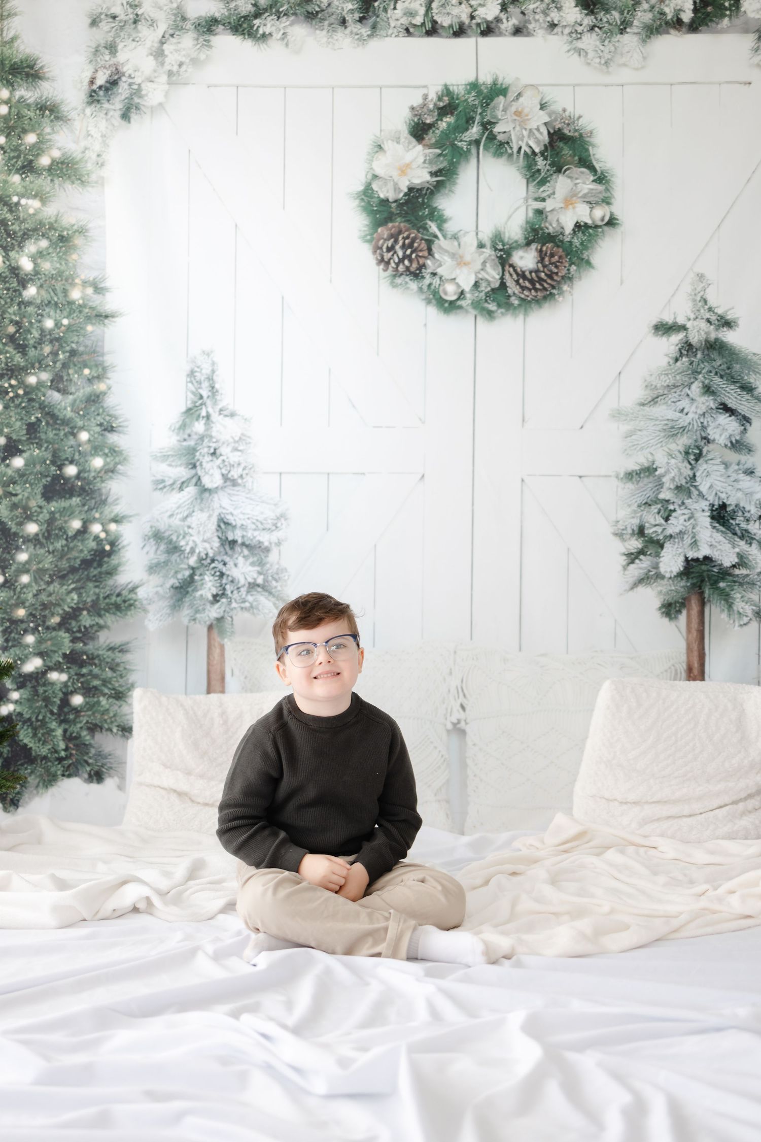 A winter portrait of a child in a dark sweater sitting on white bedding with flocked trees and wreath backdrop.