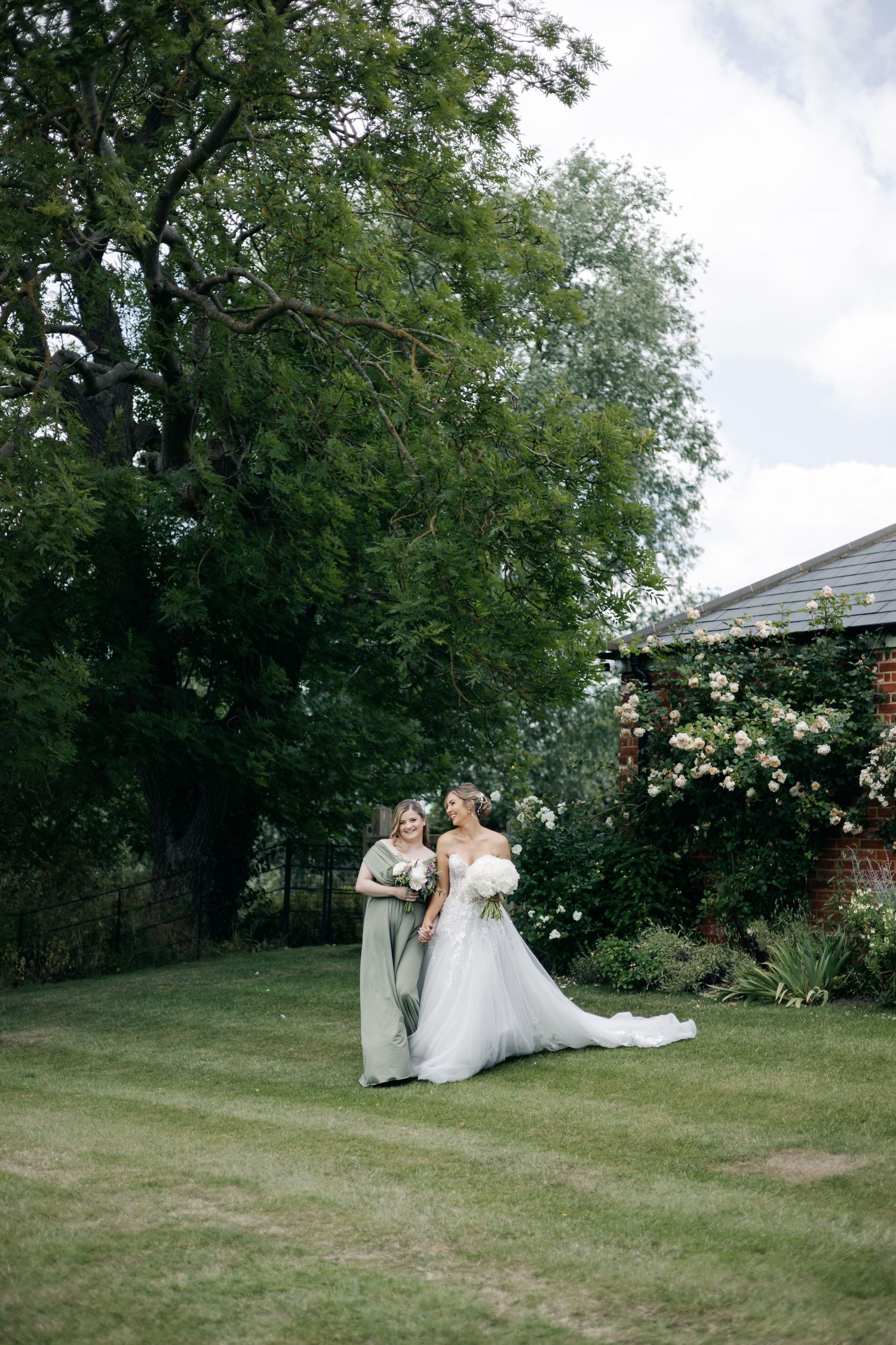 Wedding party relaxes on grassy lawn with blooming rose bushes in background.