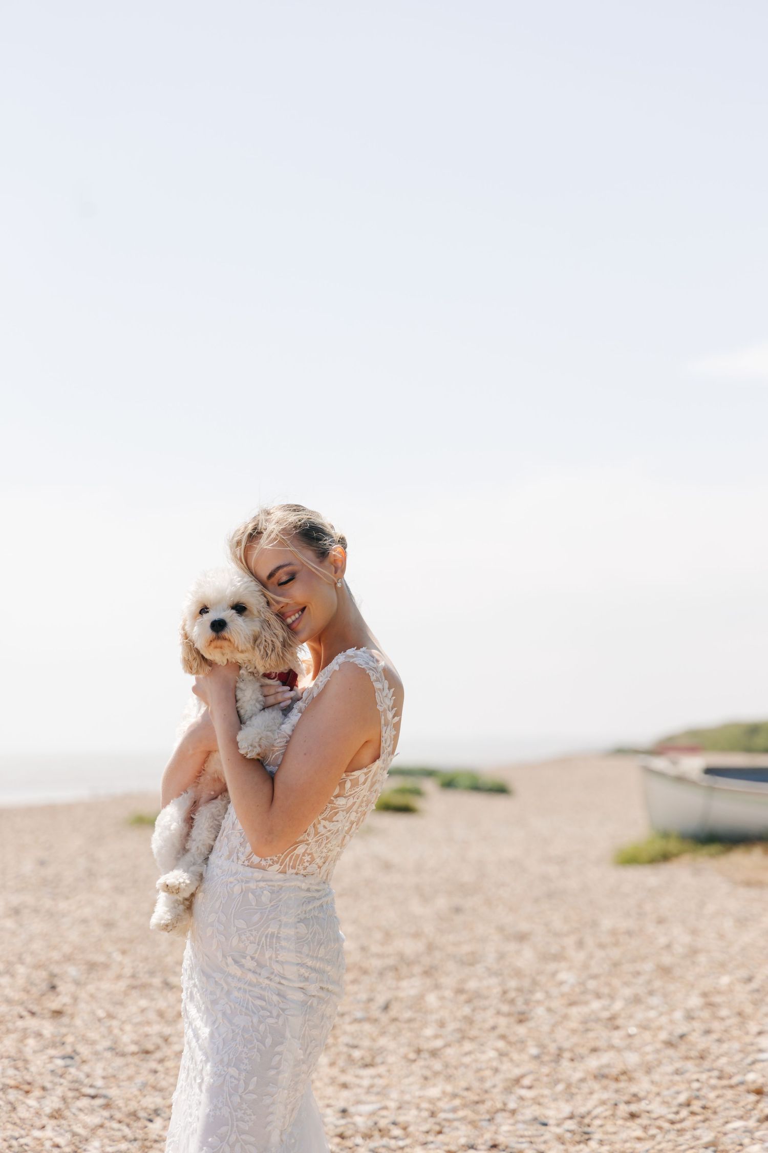 Bride in white dress embracing small white dog on a beach during wedding photoshoot.
