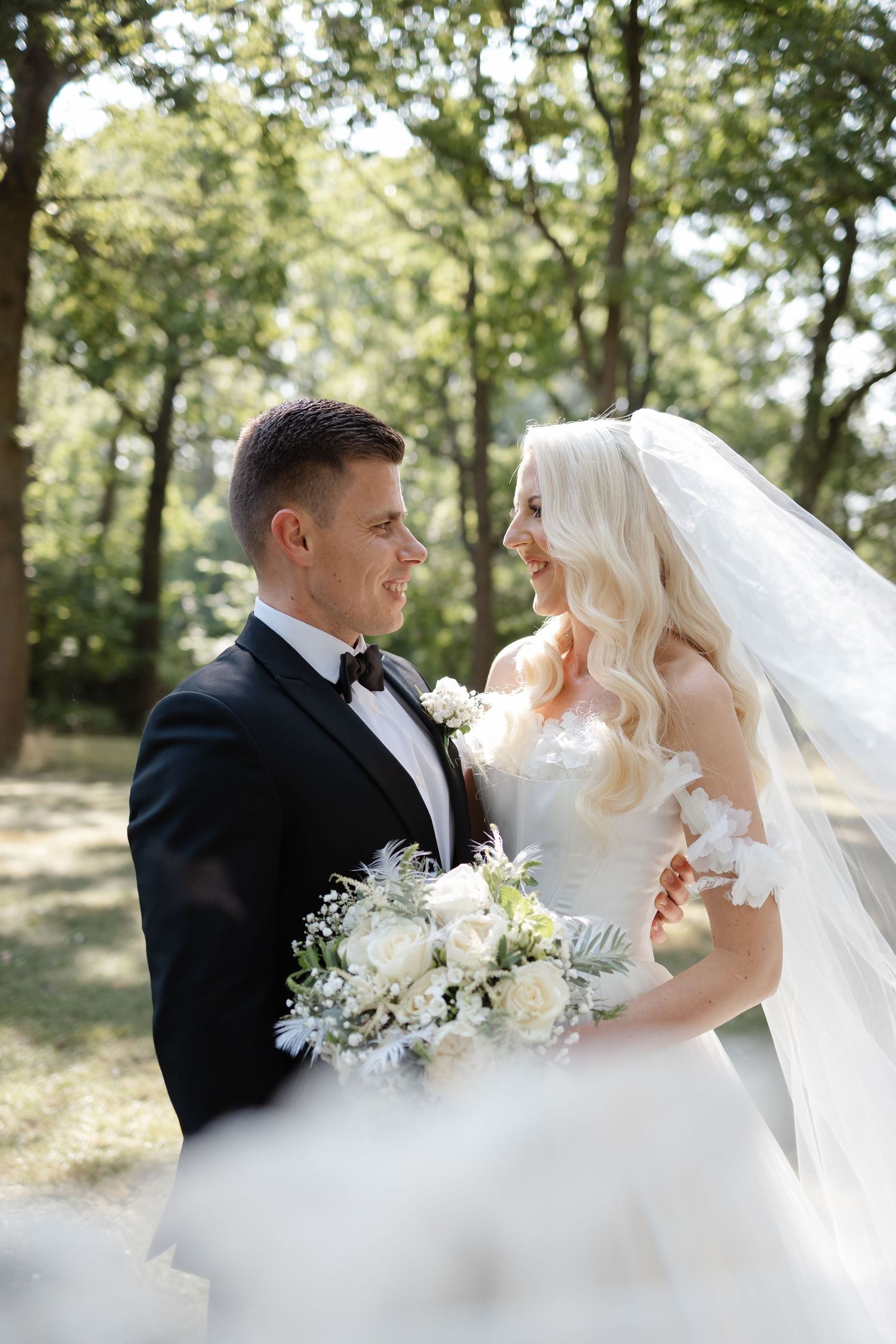 Romantic outdoor wedding portraits showing couple in sunlit garden setting with white floral bouquet.