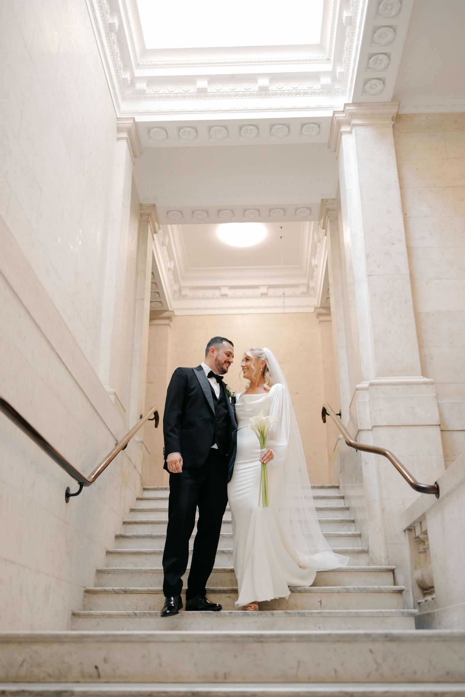 Wedding couple walking down elegant white marble stairs in an ornate building interior.