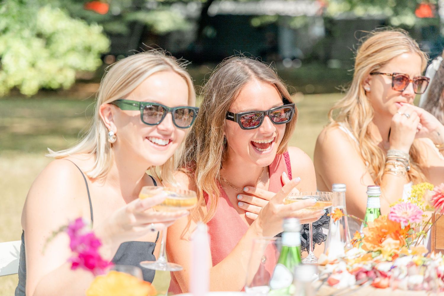 Friends sharing laughs over cocktails at sunny outdoor celebration.