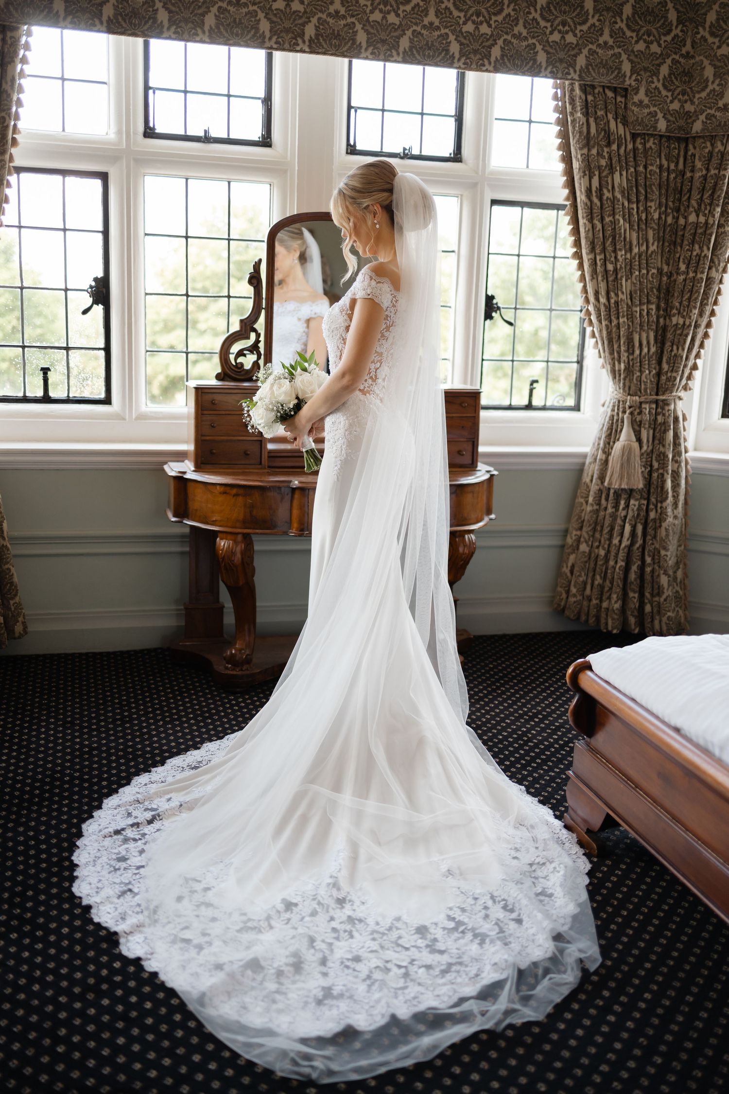 Elegant white wedding dress with lace train displayed in a sunlit room with dark carpet.