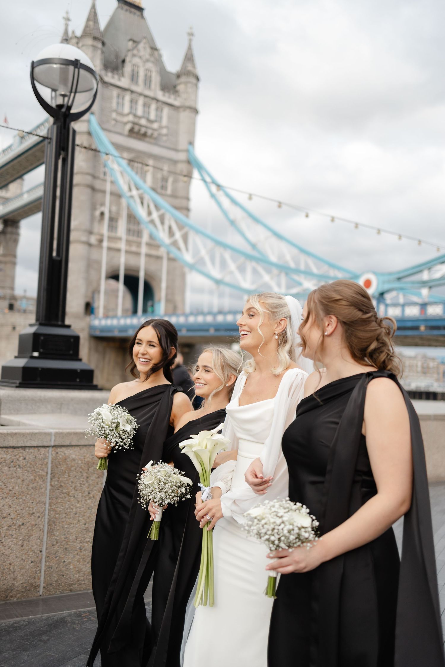 Bridesmaids in black dresses holding white bouquets pose with bride against iconic Tower Bridge in London.