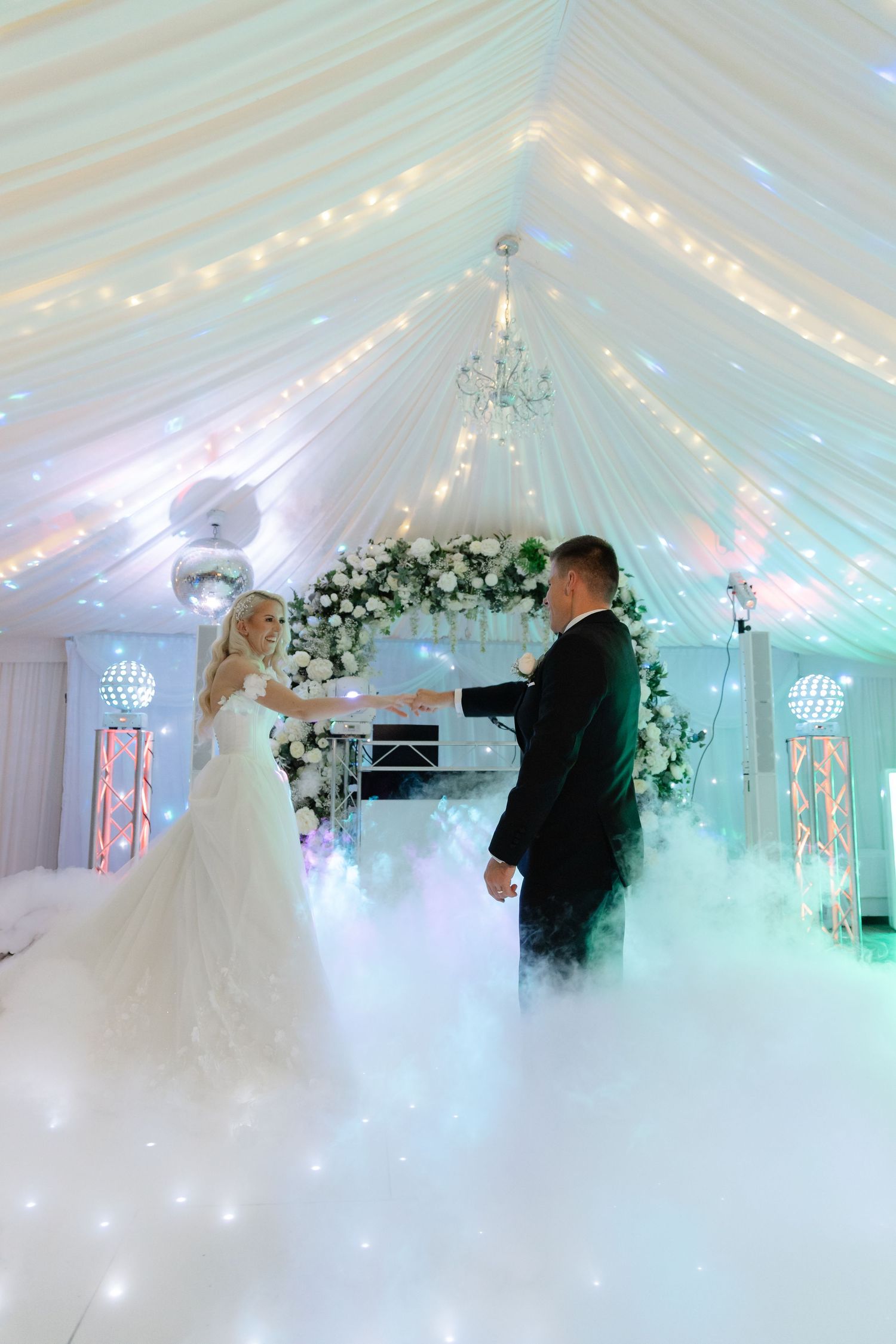 Romantic first dance in a beautifully lit white marquee with twinkling lights and floral arrangements.