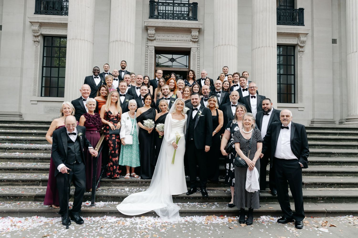 Large wedding party gathered on grand stone steps for formal group photo.