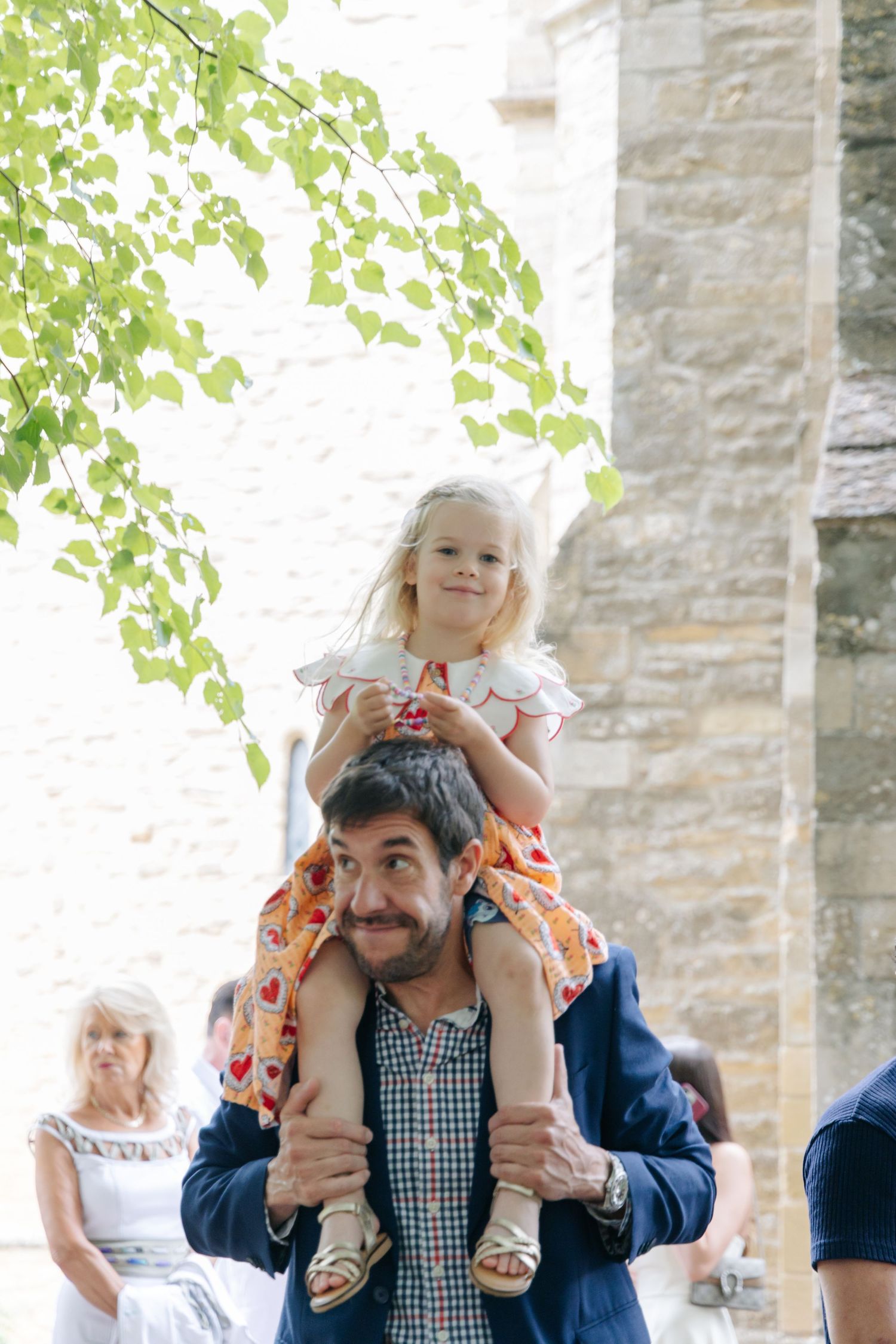 A playful moment outside a stone building with someone carrying a child on their shoulders.