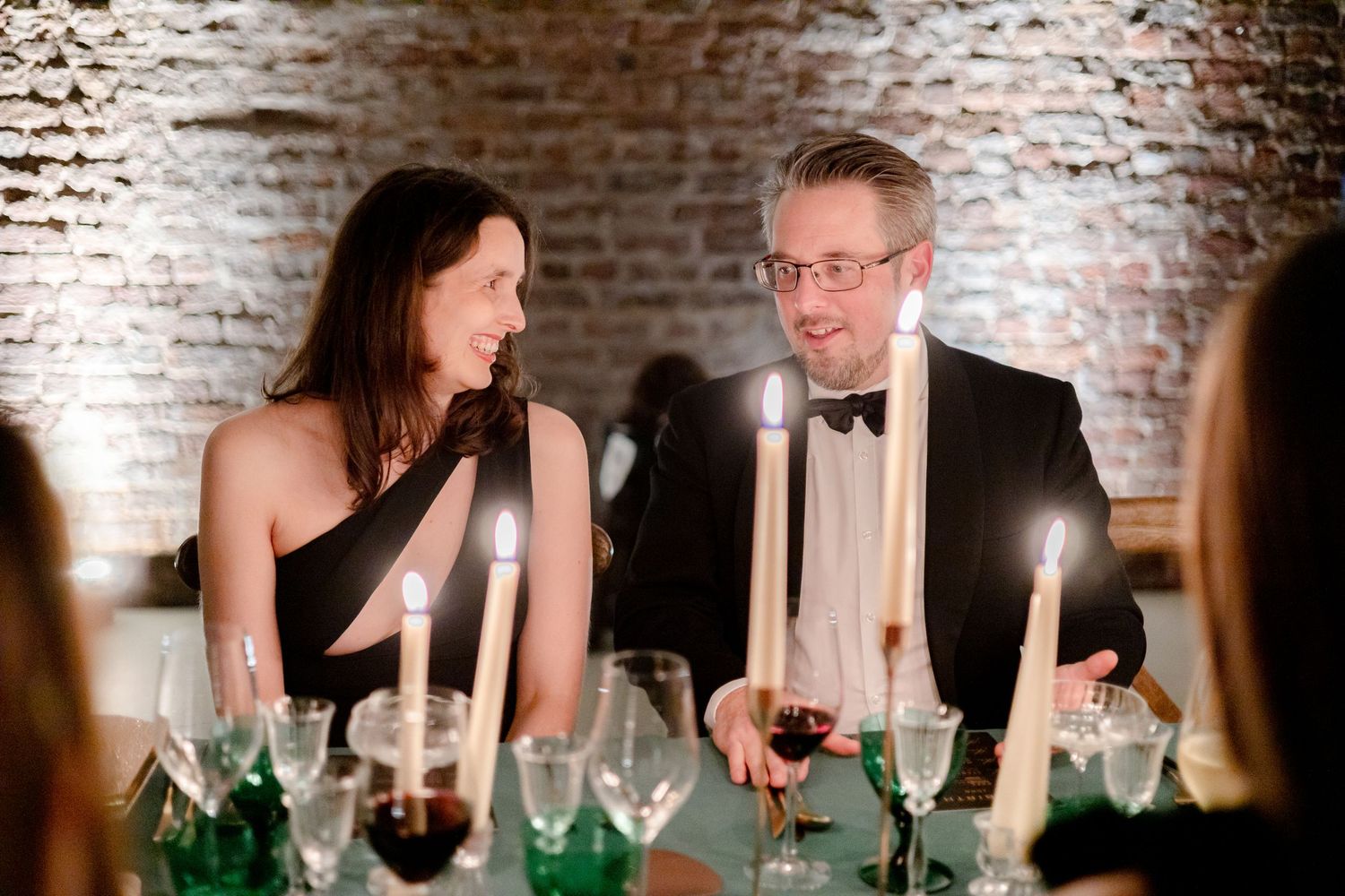 Elegant dinner party guests conversing at a candlelit table with wine glasses and formal attire.