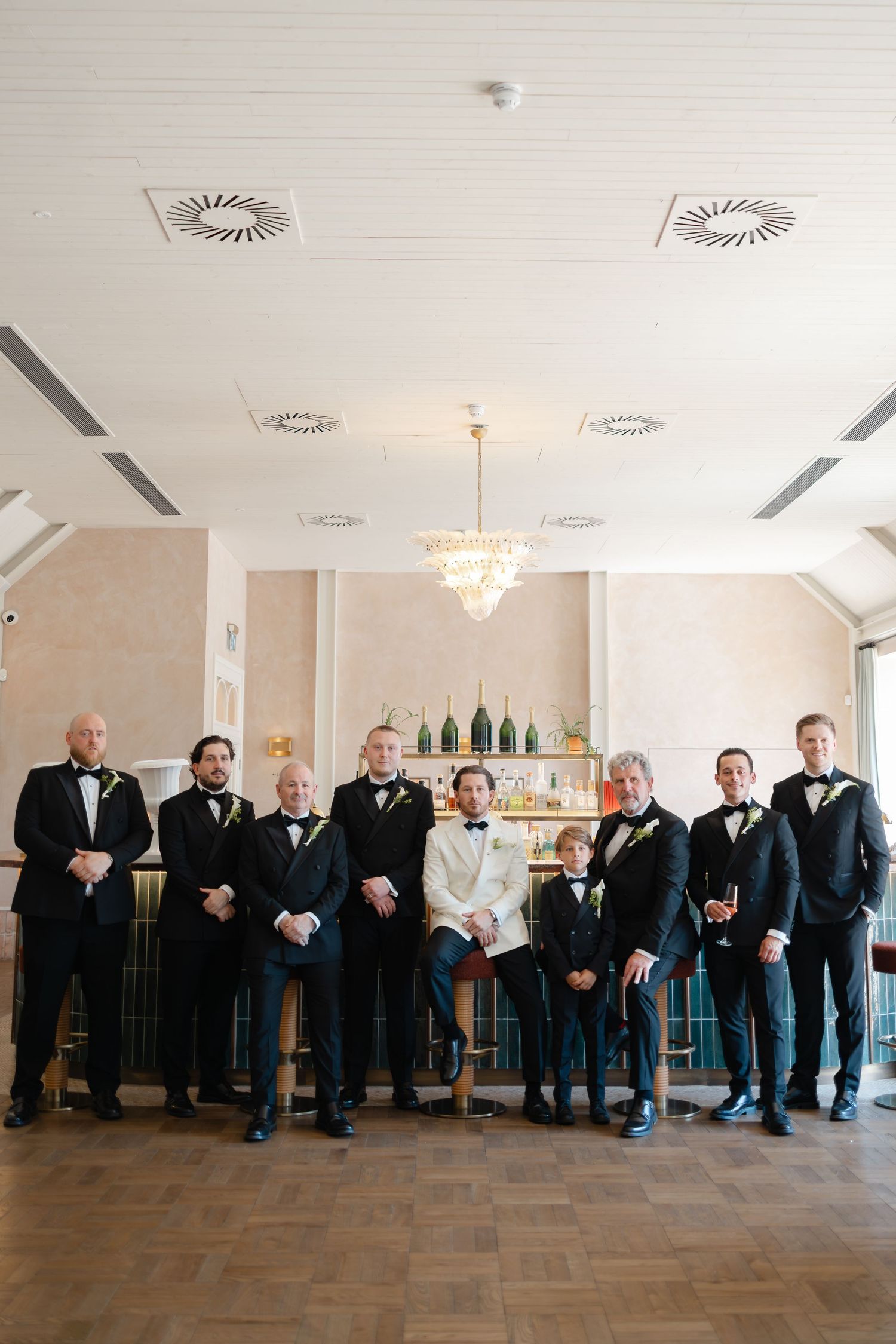 Groom in white tuxedo jacket stands with groomsmen in black tuxedos before an elegant bar backdrop.