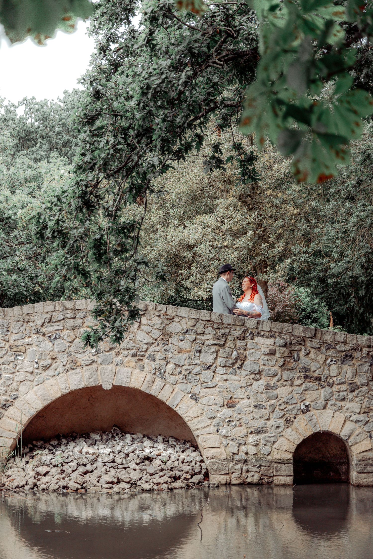 A couple sitting on a stone bridge over water surrounded by lush greenery.
