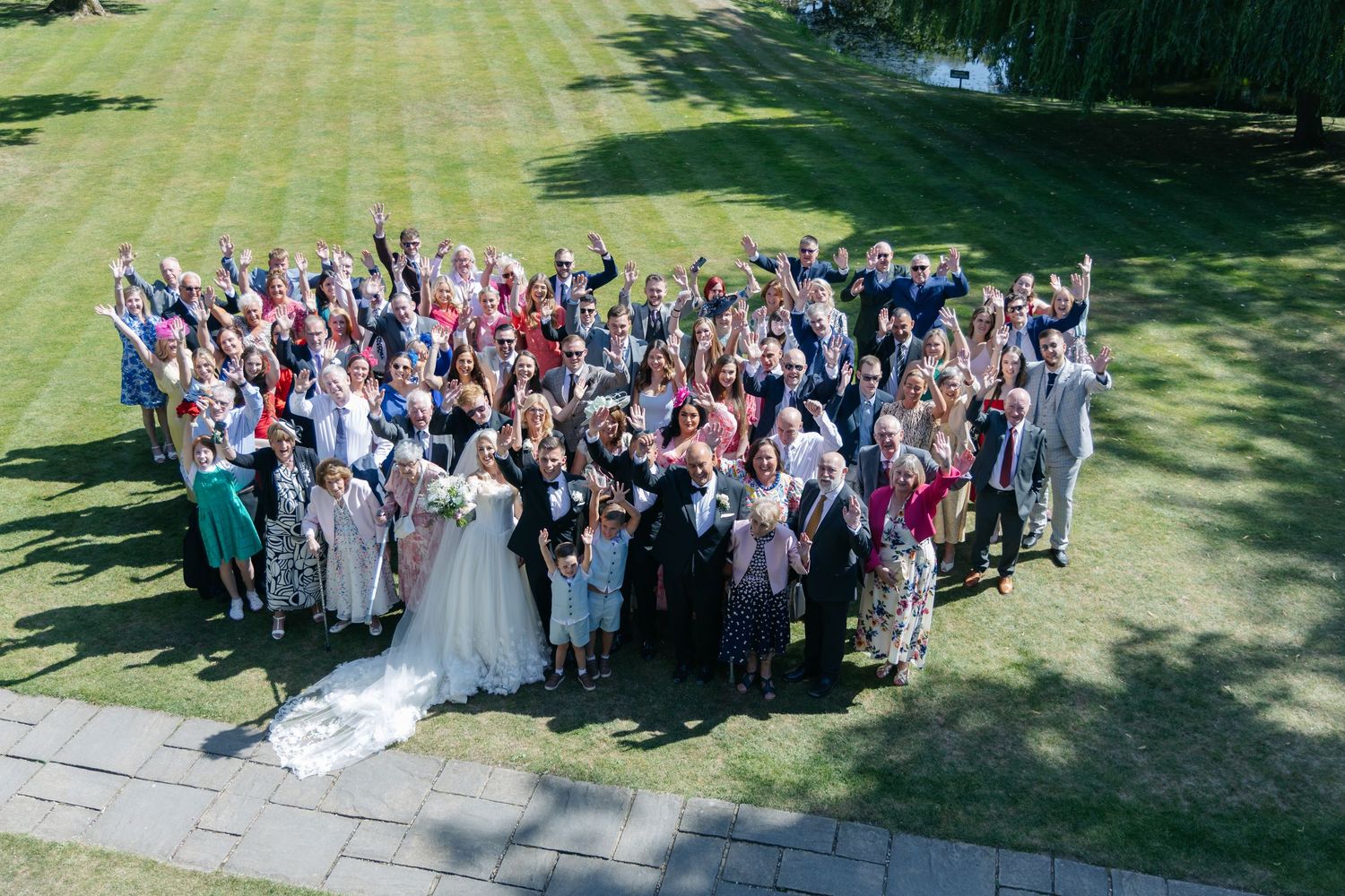Aerial view of wedding party and guests forming large group photo on manicured lawn.