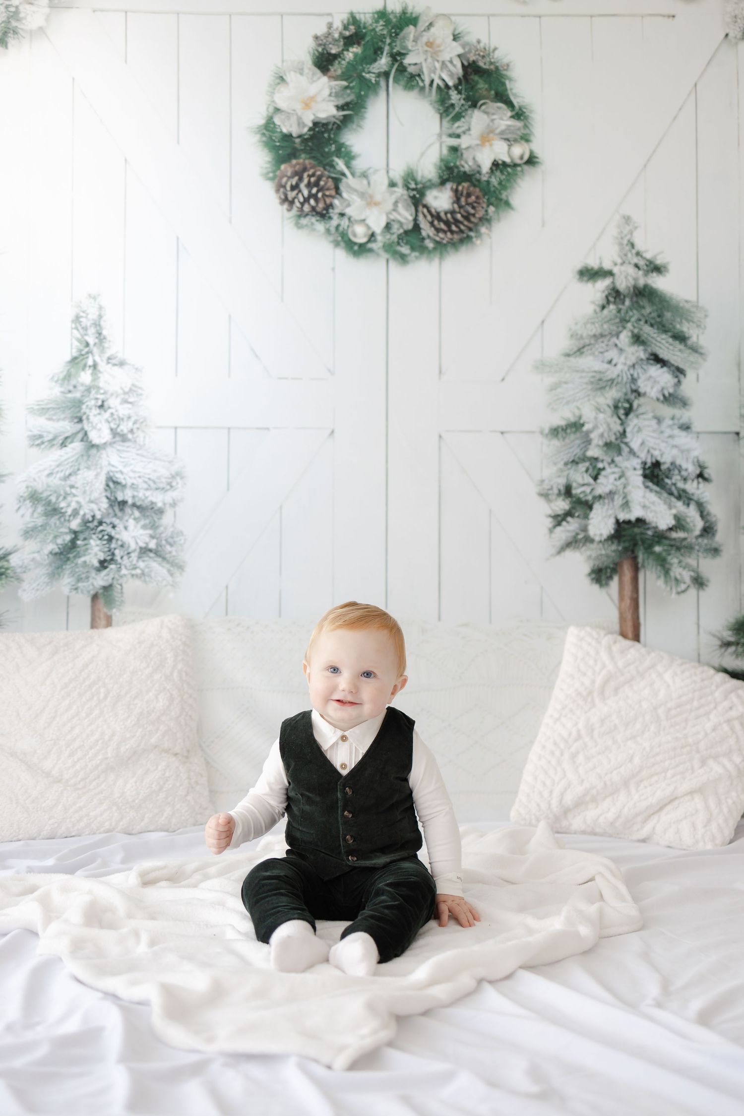 Small child in black formal wear sits on white bedding with winter decorations in background.