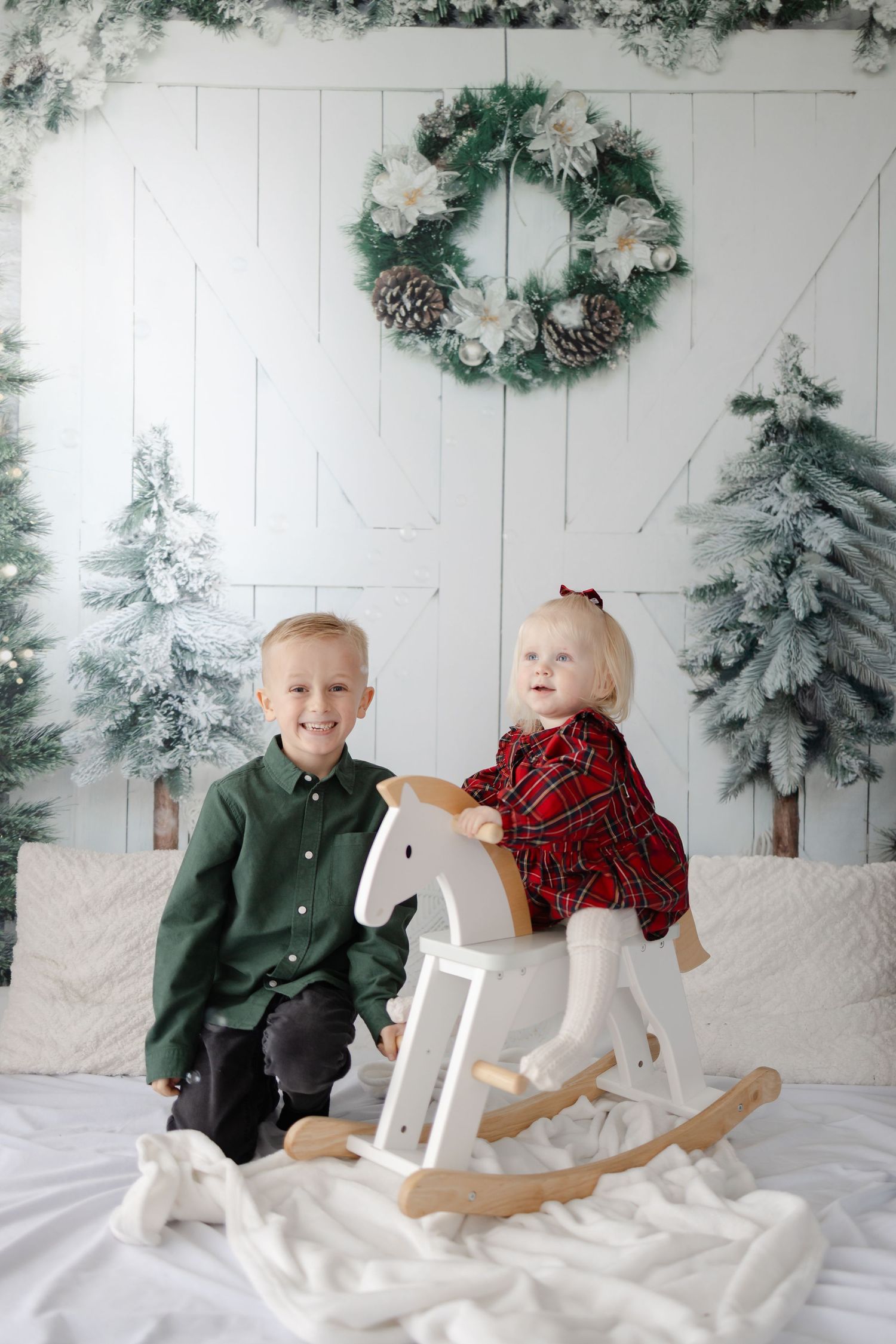 Two children in formal attire play with a white rocking horse against a winter-themed backdrop.