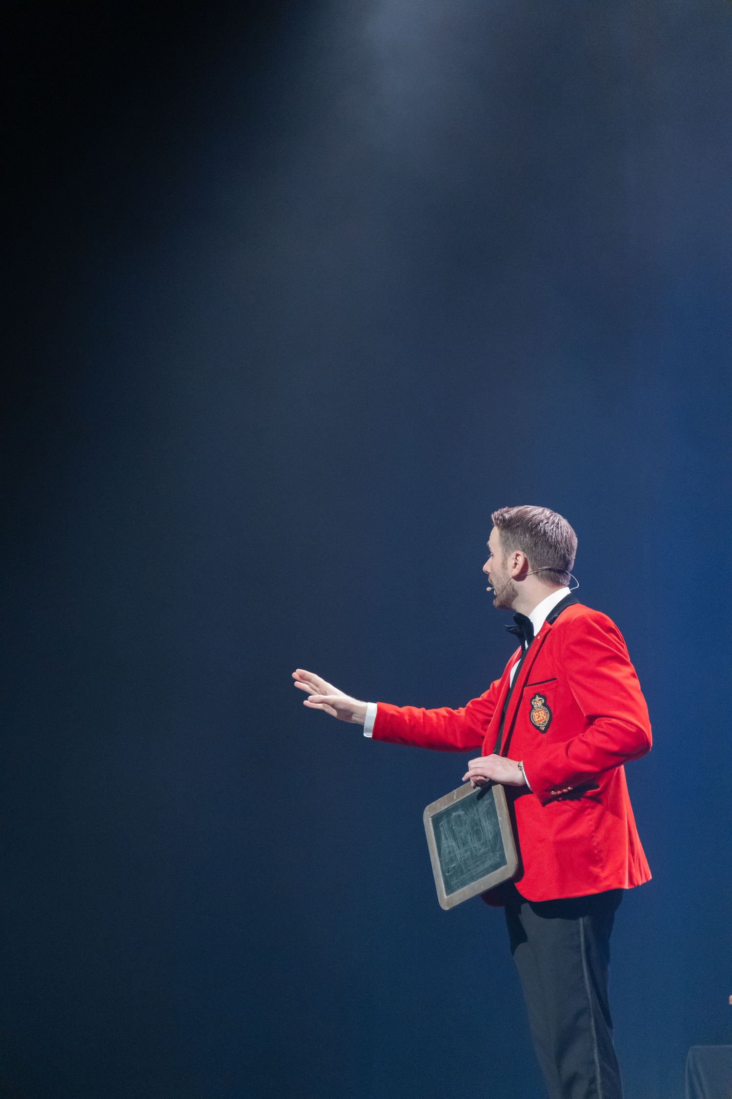 Dramatic performance scene with person in red jacket gesturing on stage.