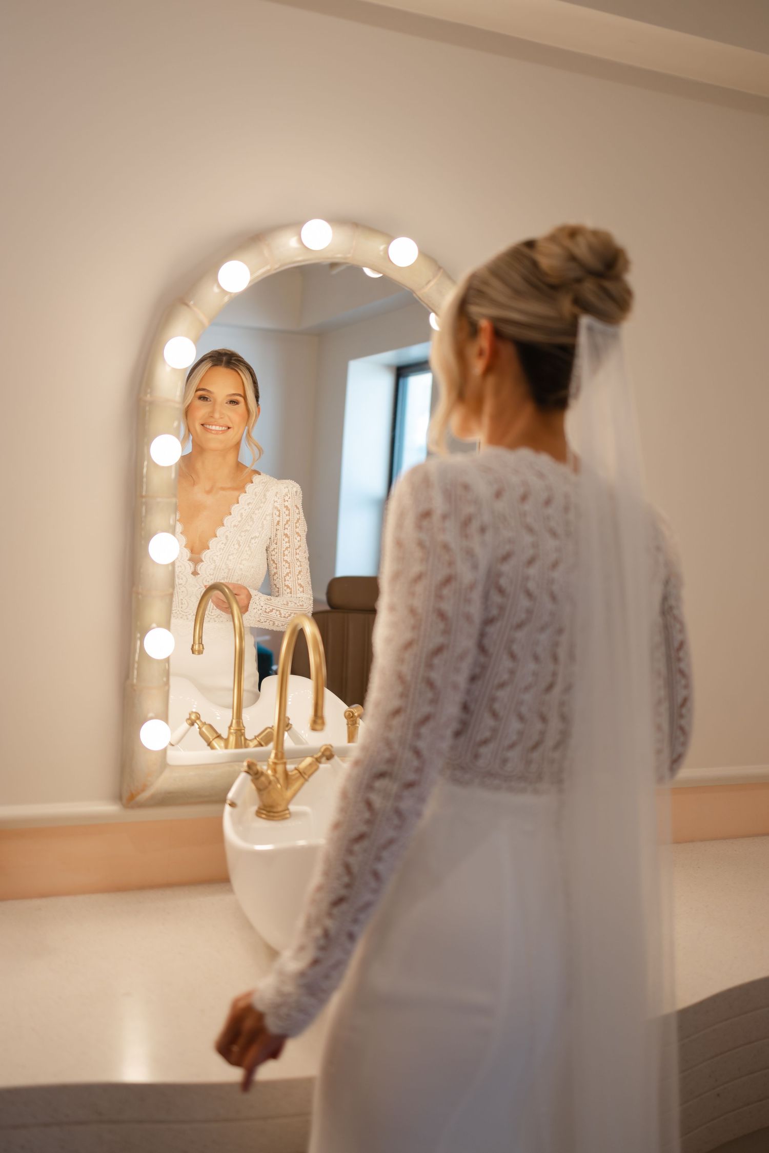Smiling bride in lace wedding dress admires her reflection in a Hollywood mirror with bulb lights.