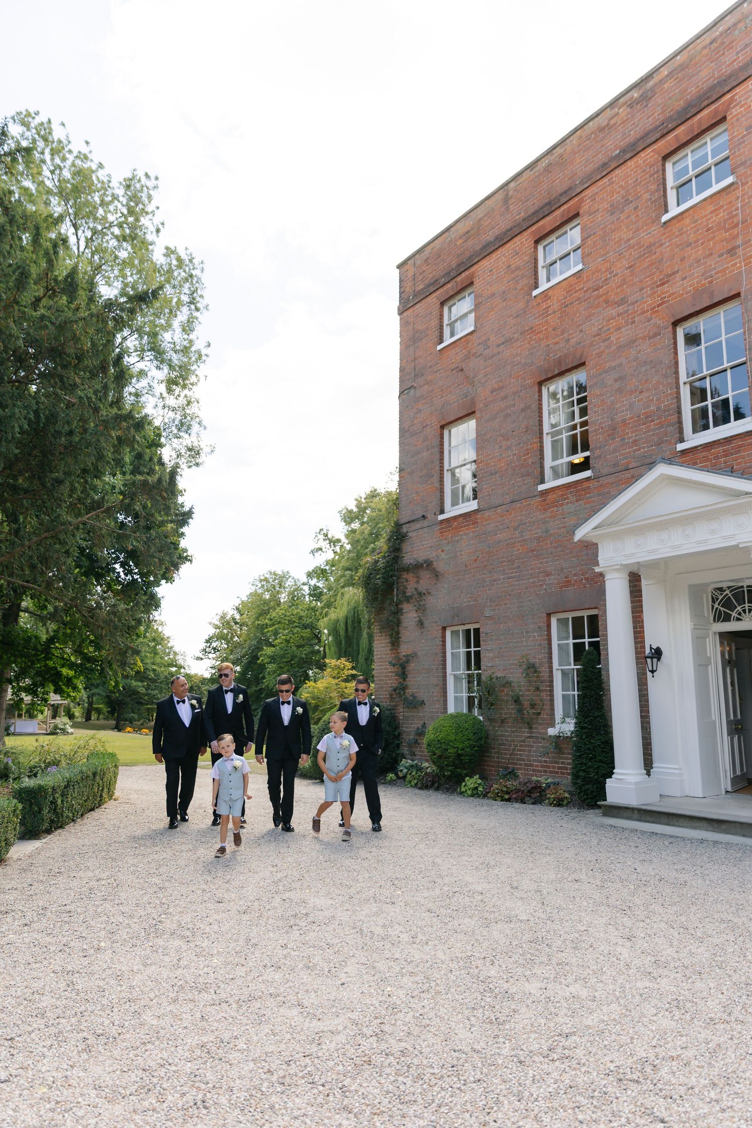 Elegant brick manor house with guests walking on gravel path surrounded by greenery.