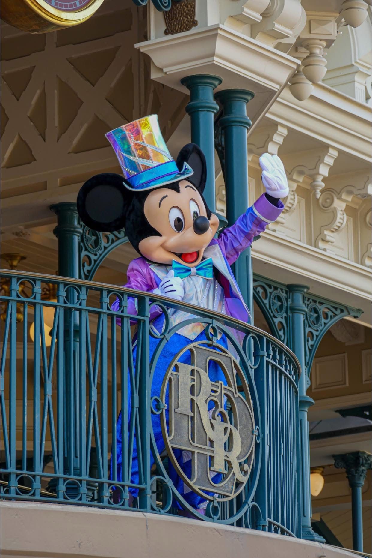 Mickey Mouse character in a blue and white conductor uniform waves from a balcony at Disney theme park.