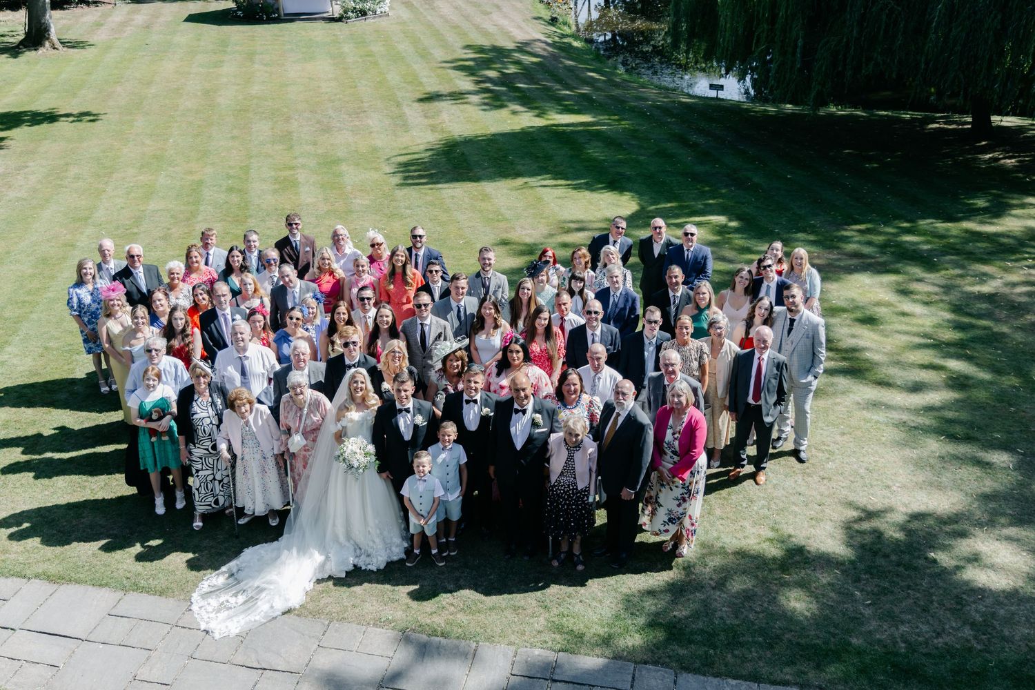 Large group wedding photo taken outdoors on a sunny day with guests arranged in rows.