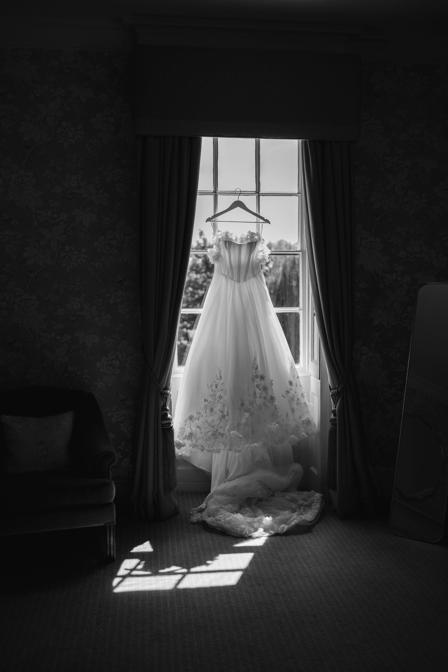 A white wedding dress hangs elegantly in front of a sunlit window creating a dramatic silhouette.