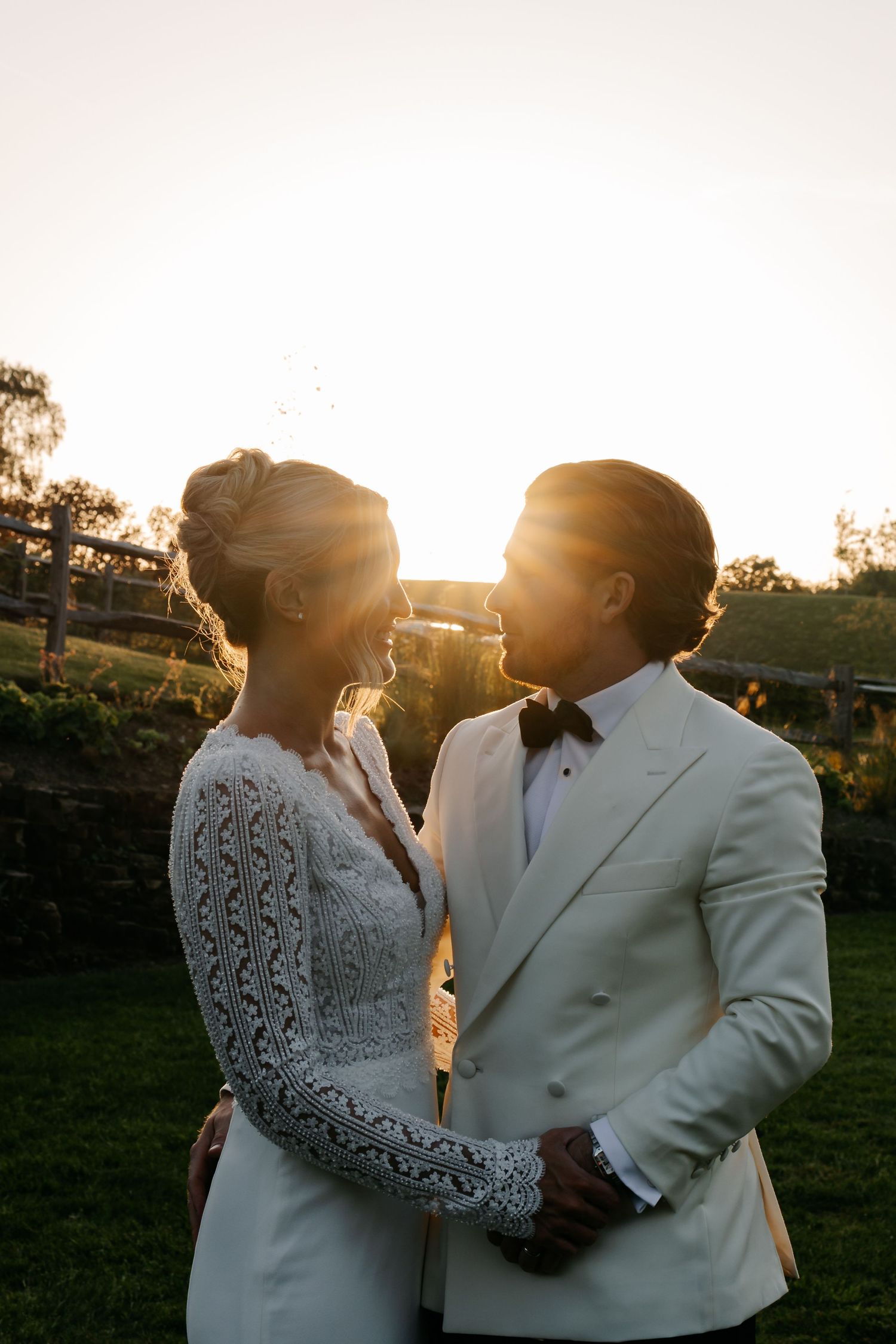 Bride and groom share a romantic sunset moment, she in lace gown, he in white tuxedo, golden light glowing behind them.