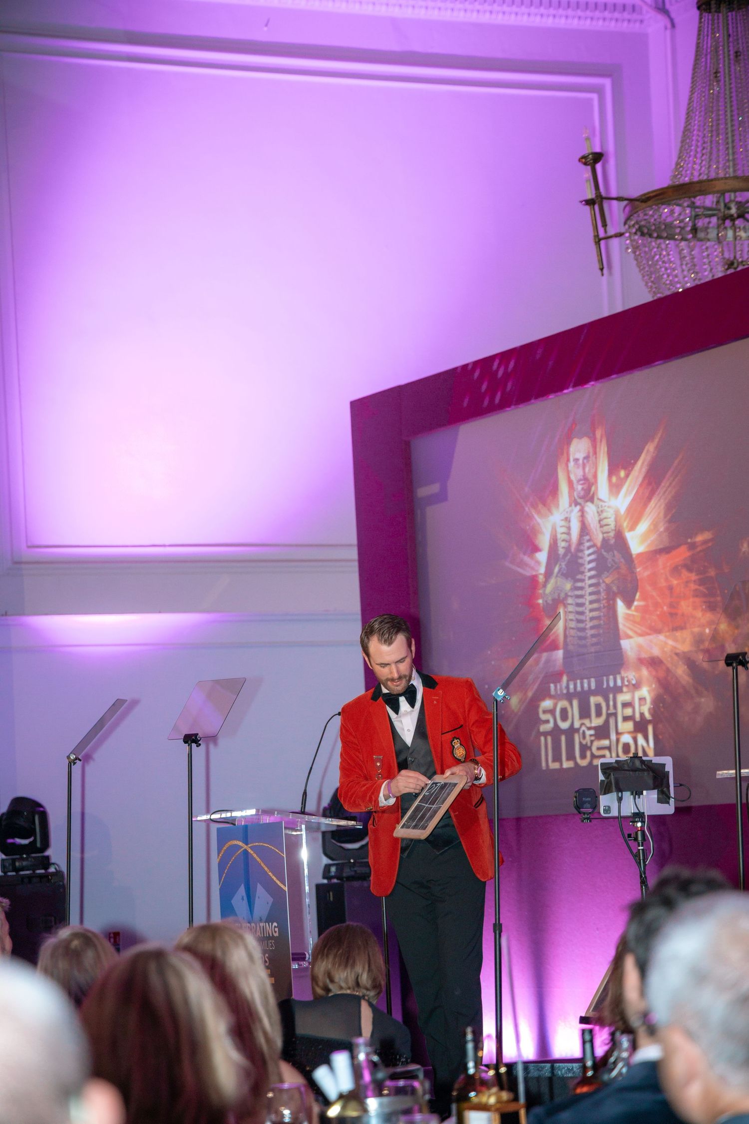 Speaker at podium during formal event with red jacket and projection screen behind.
