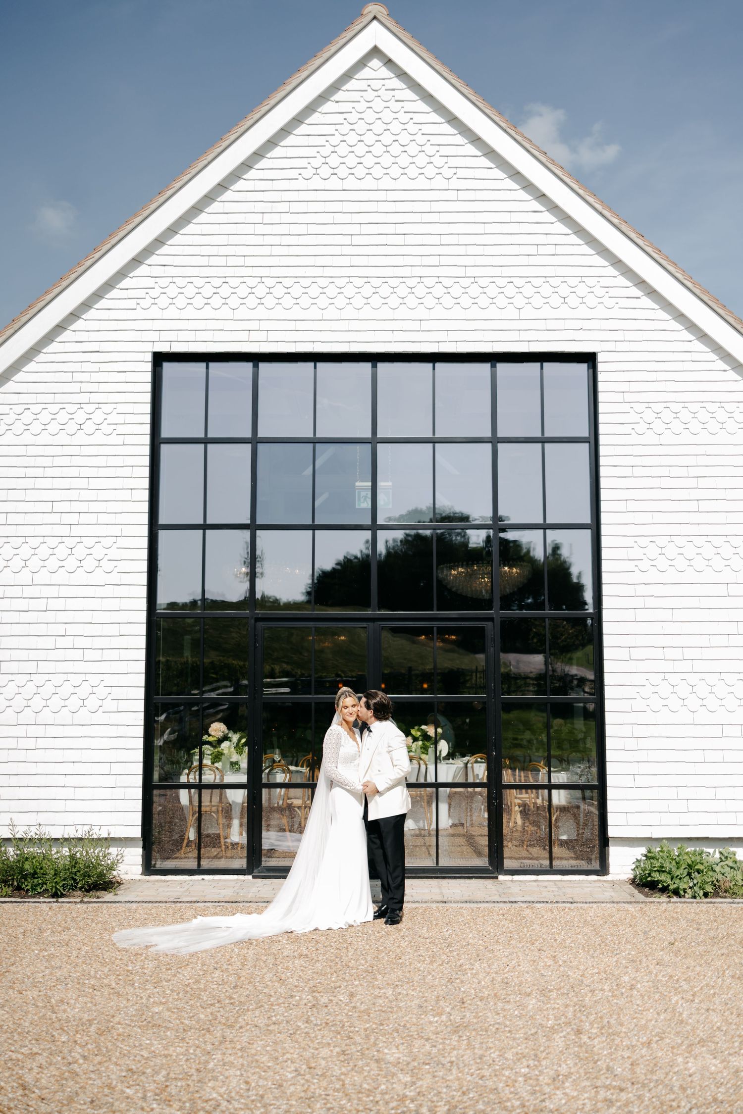 Bride and groom embracing in front of a white barn wedding venue with large black-framed windows under a blue sky.