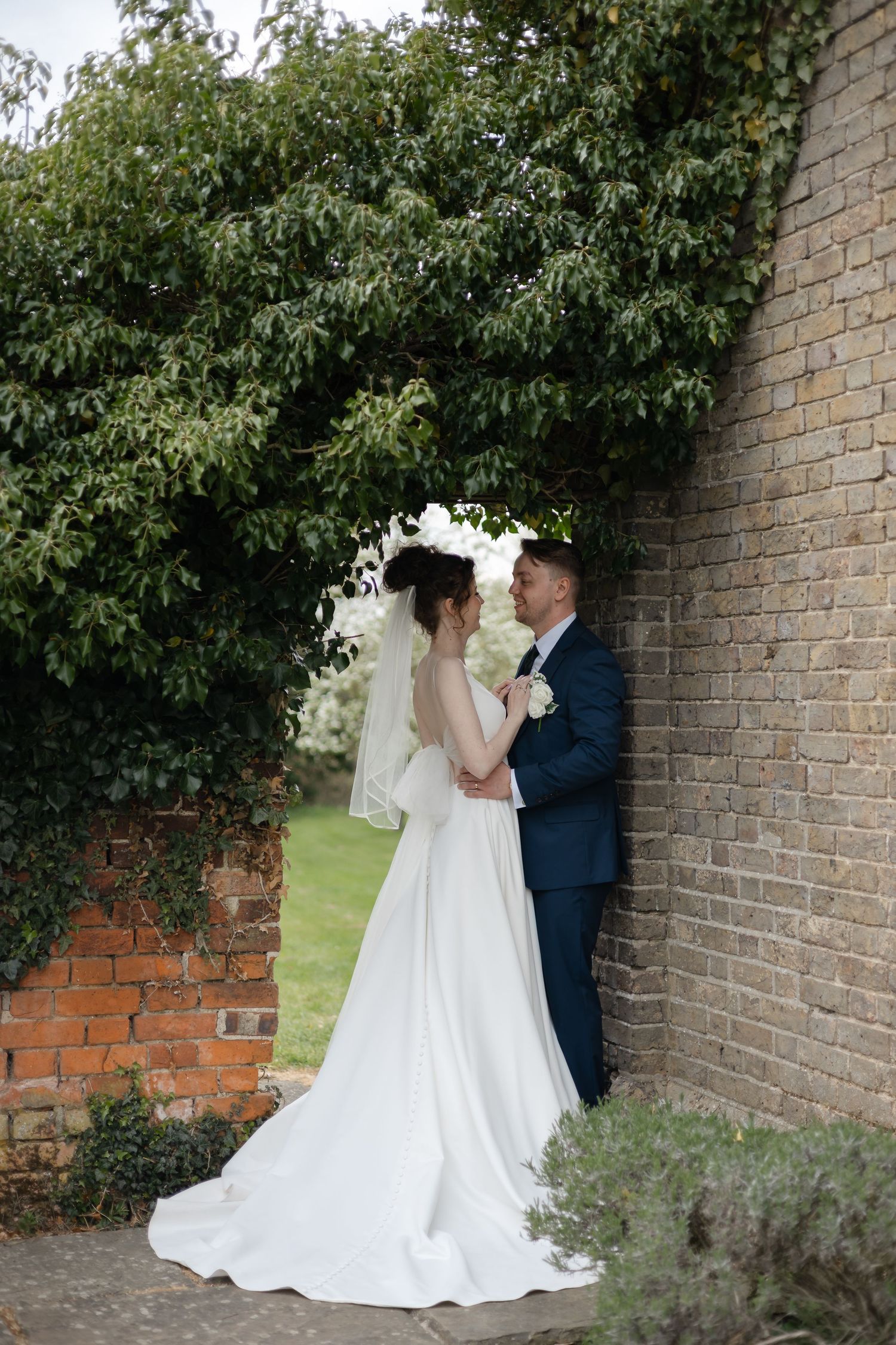 Newlyweds share intimate moments against a brick wall backdrop surrounded by lush foliage.