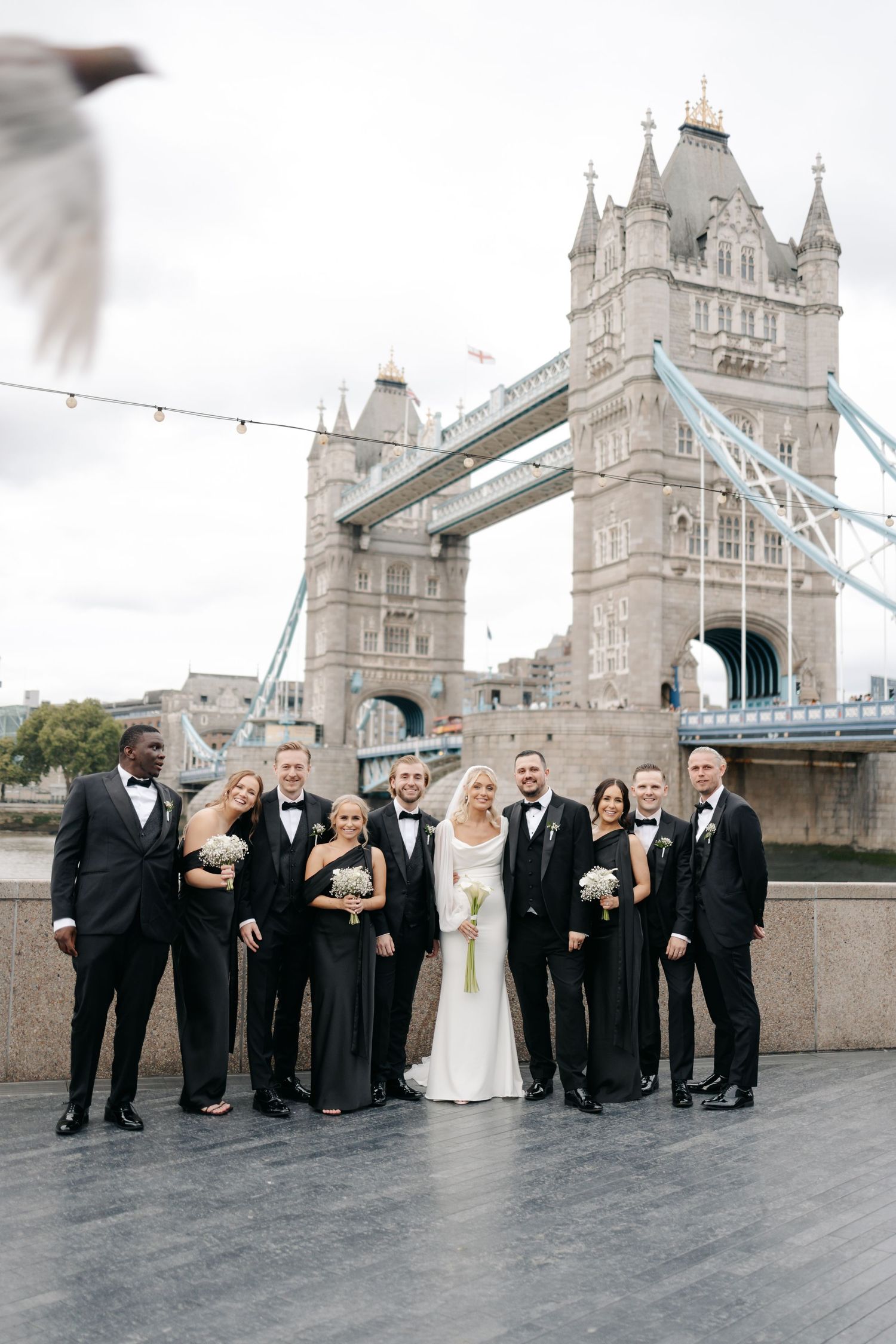 Wedding party in black and white attire poses for group photos with Tower Bridge backdrop.
