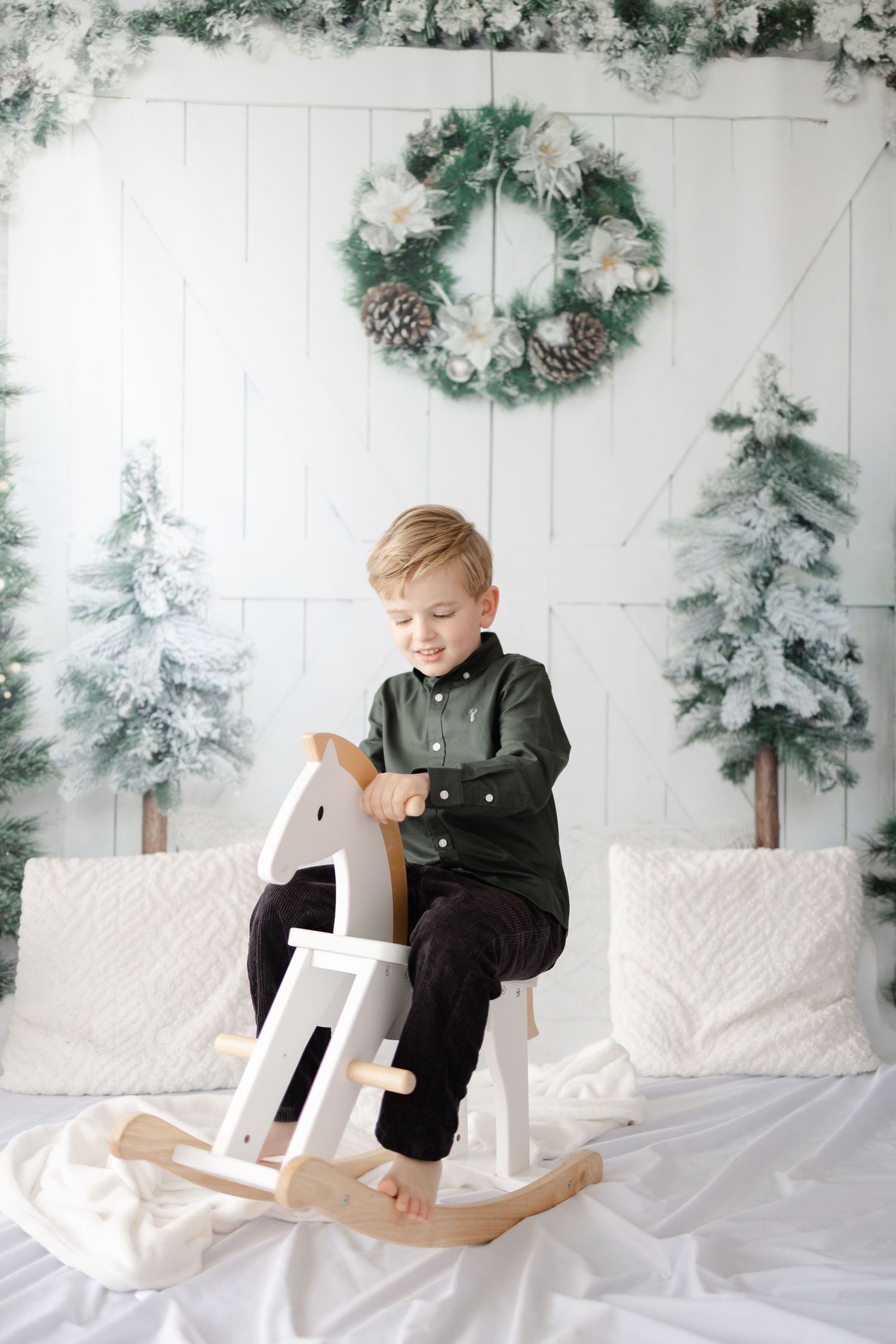 Child playing on white wooden rocking horse with snowy Christmas trees in background.