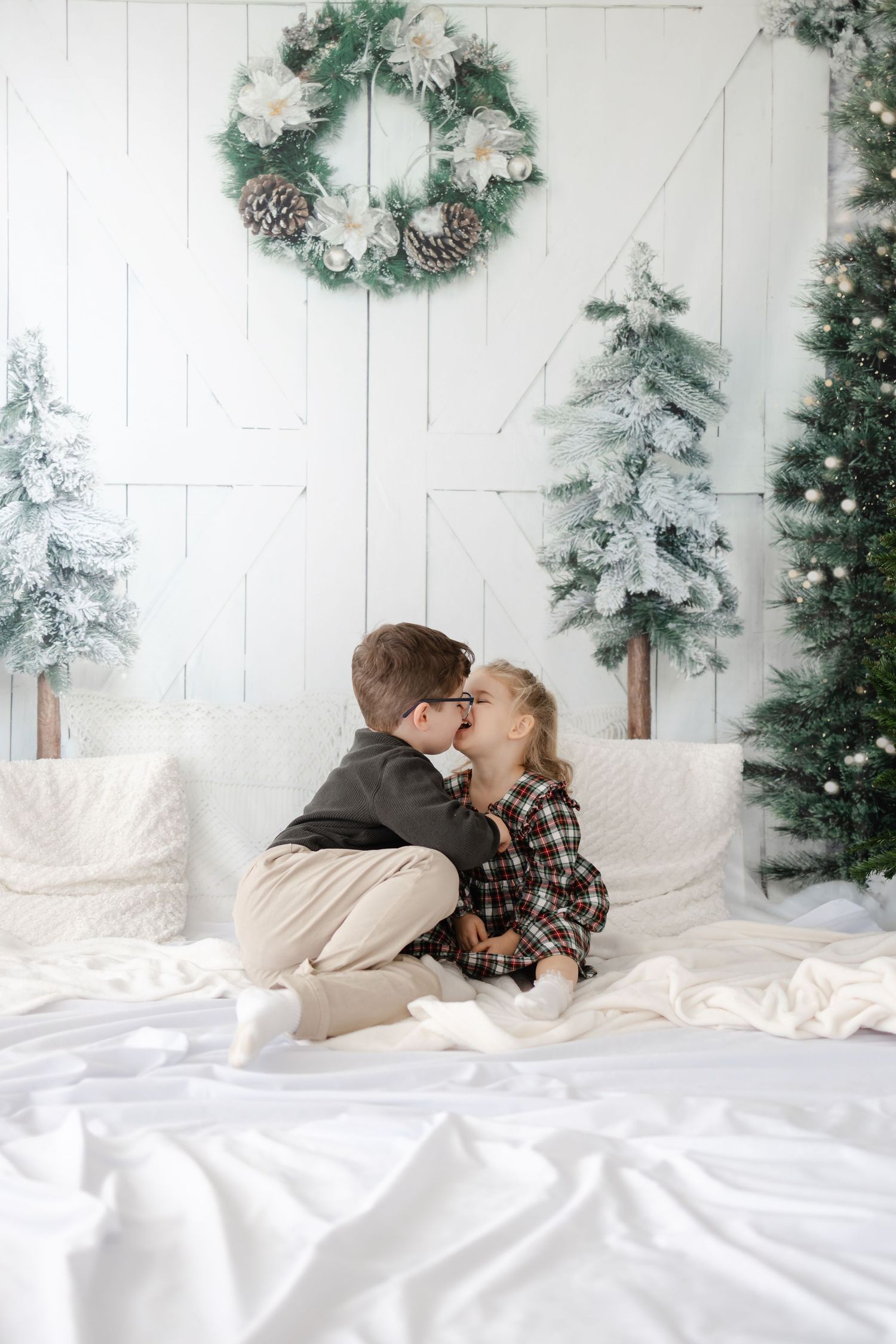 Two children share a sweet moment on white bedding with holiday decorations and flocked trees in the background.