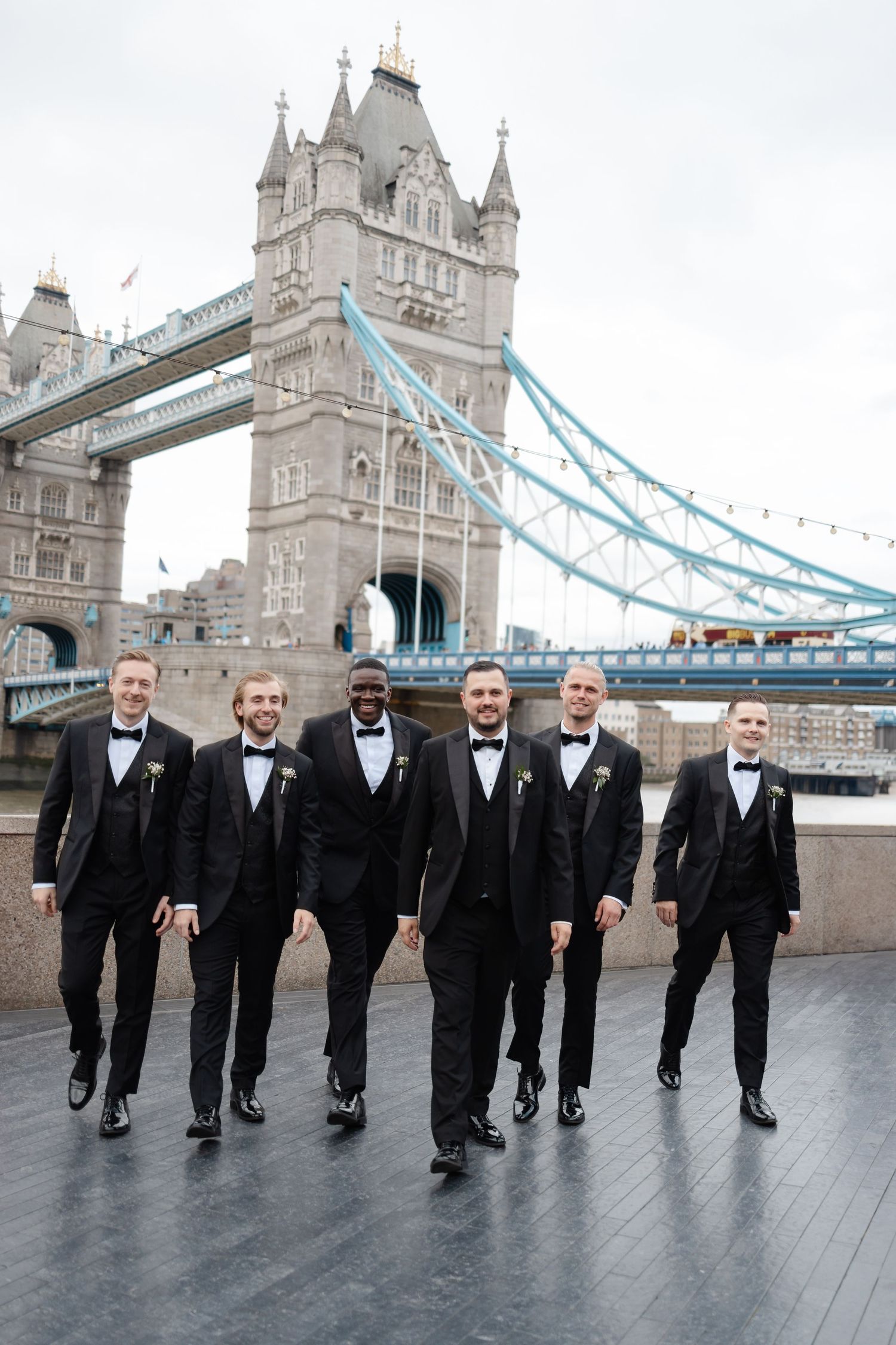Groomsmen in black tuxedos walking in line with Tower Bridge visible behind them.