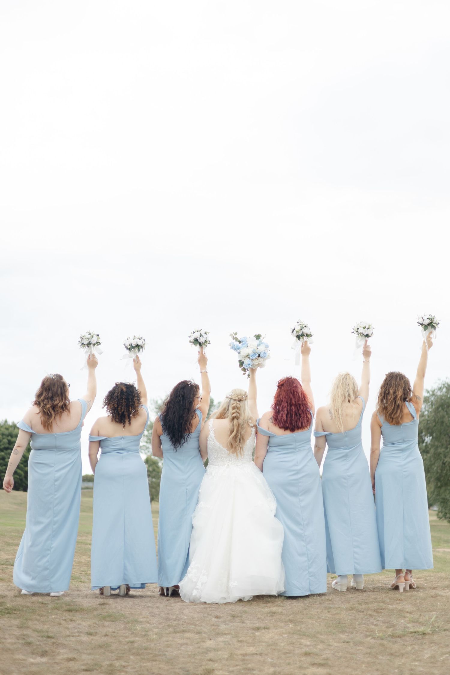 Wedding party in light blue bridesmaid dresses and white bridal gown pose with raised bouquets from behind.