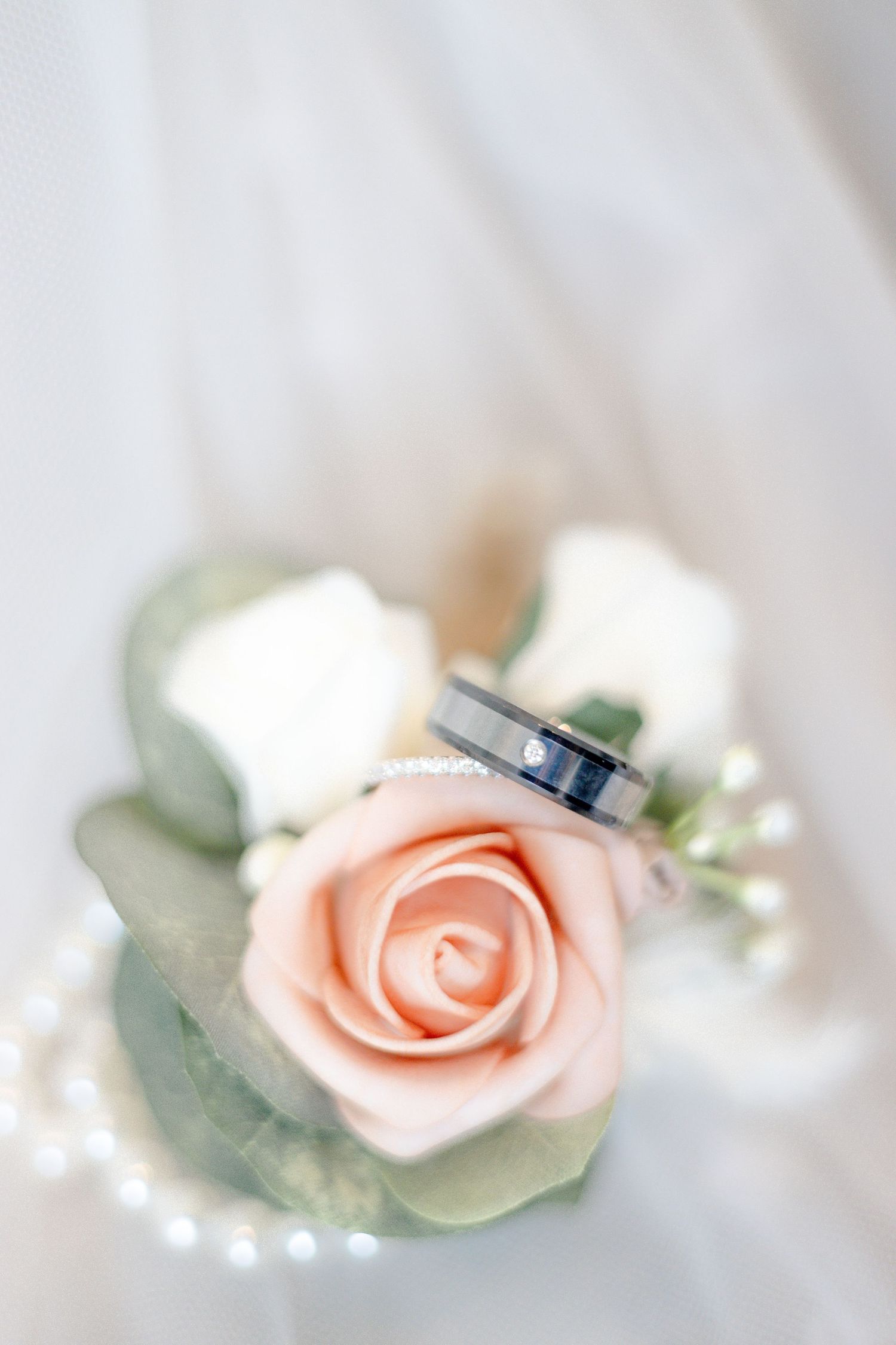 A delicate peach rose with wedding rings and pearls arranged on a soft white background.