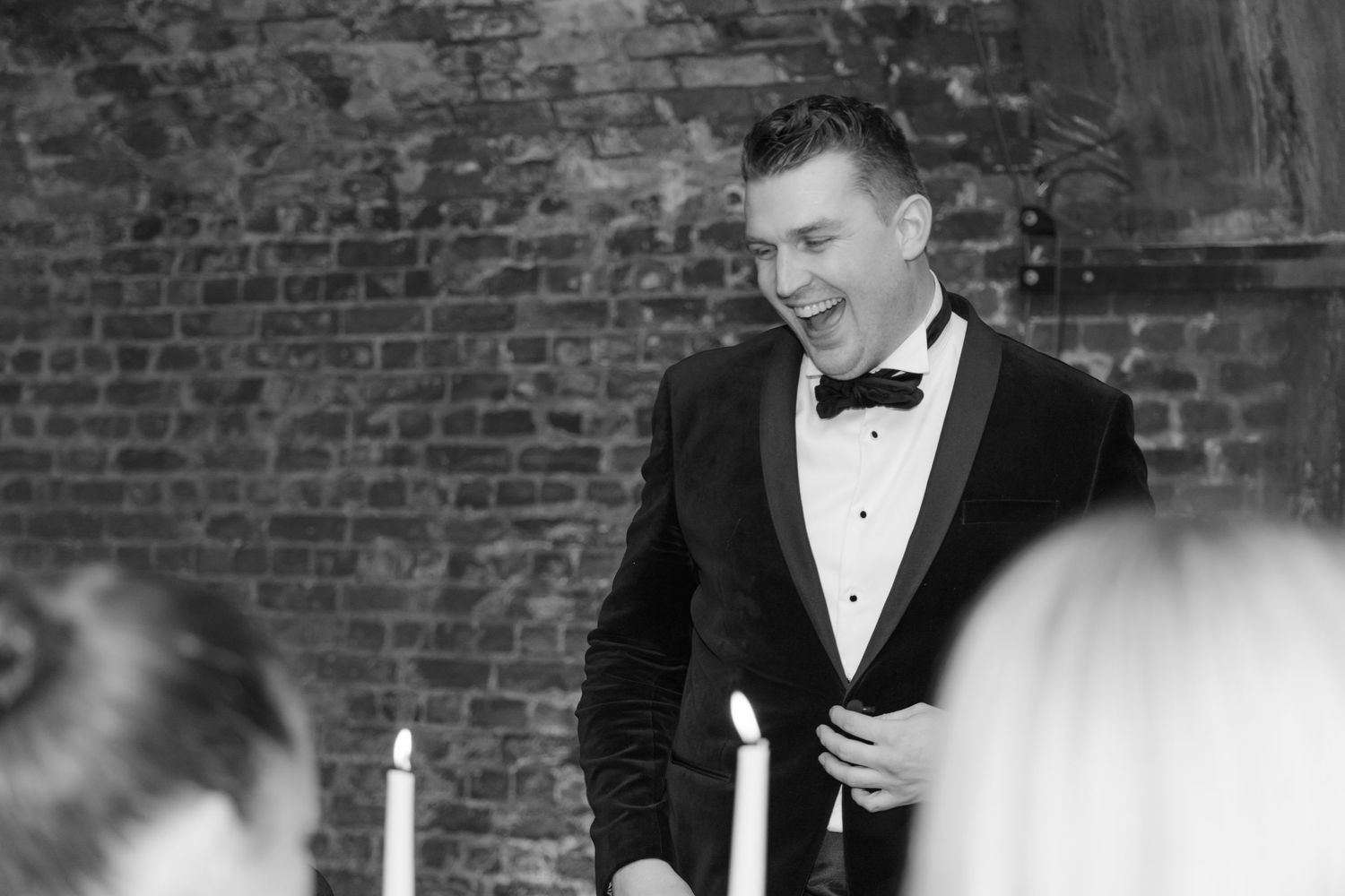 Person in black tuxedo smiling during formal dinner event with exposed brick wall backdrop.