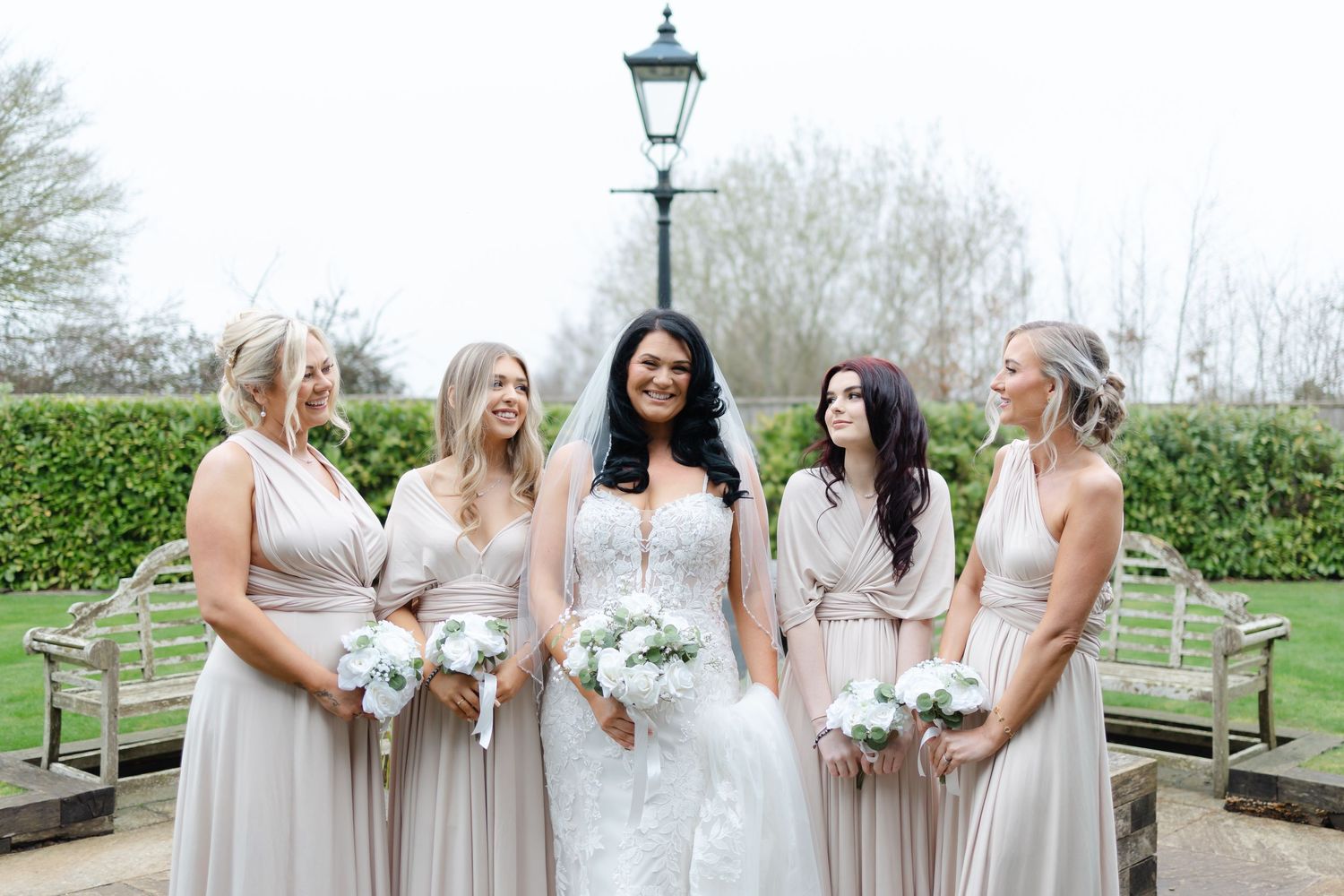 Bridesmaids in light colored dresses pose with bouquets in outdoor garden setting.