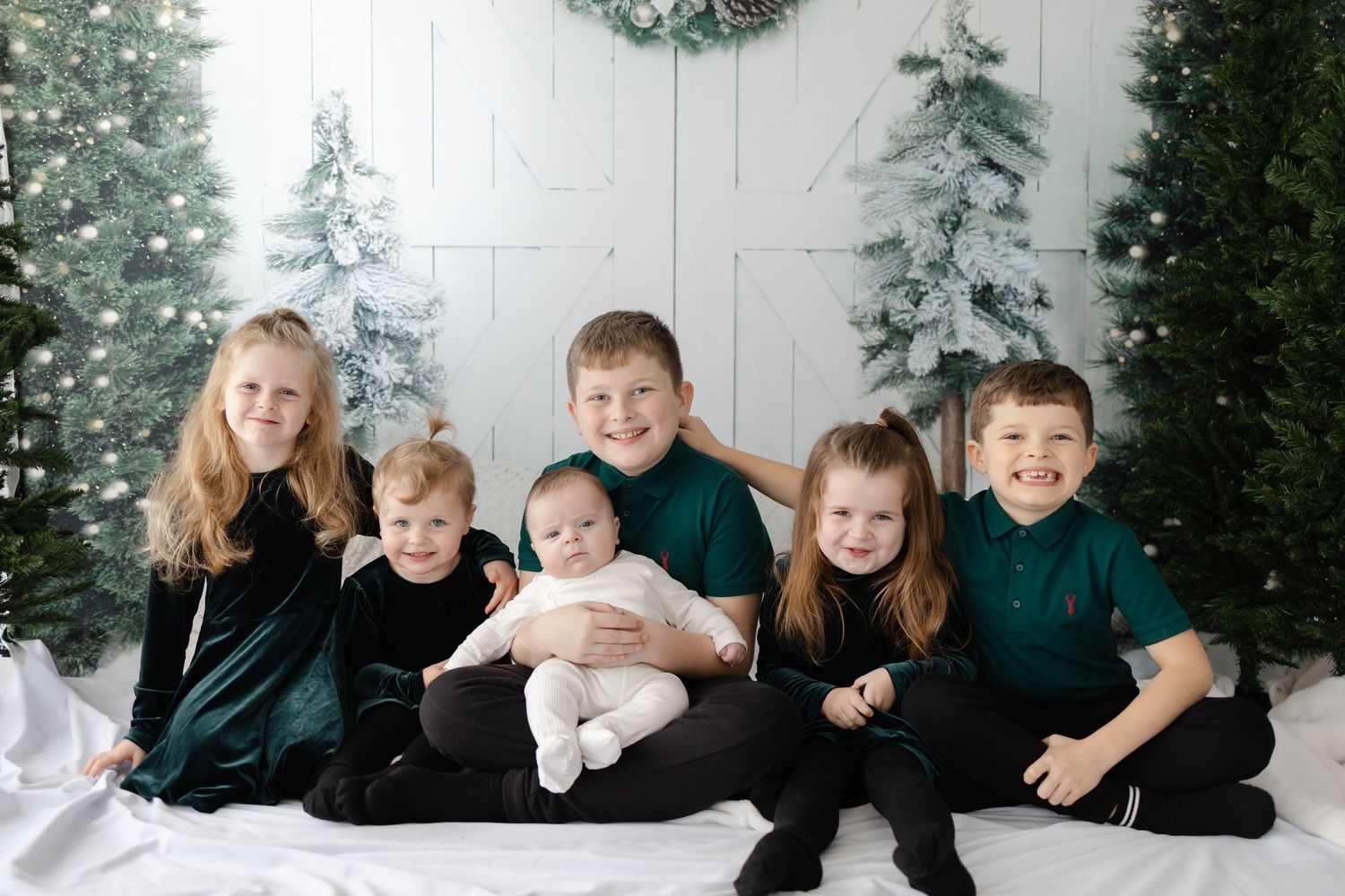Siblings in black outfits pose together against a winter backdrop with evergreen trees and white decor.