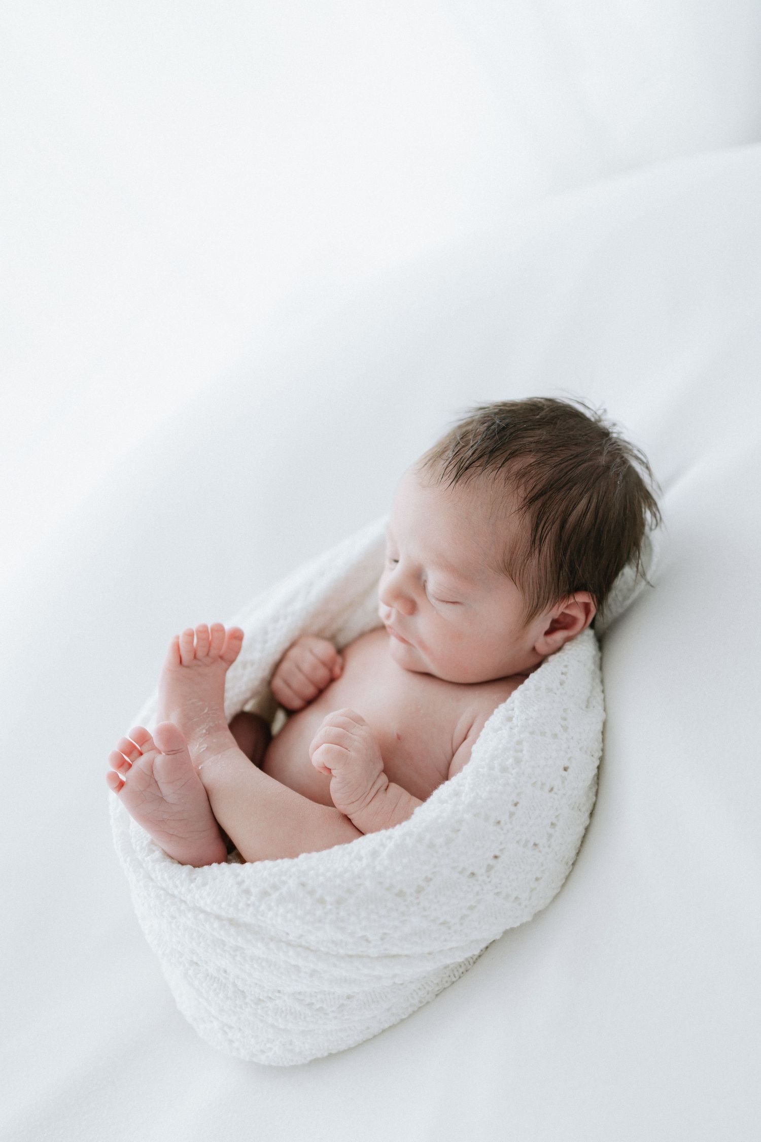 Peaceful newborn baby sleeping wrapped in a white knit blanket on a soft white background.