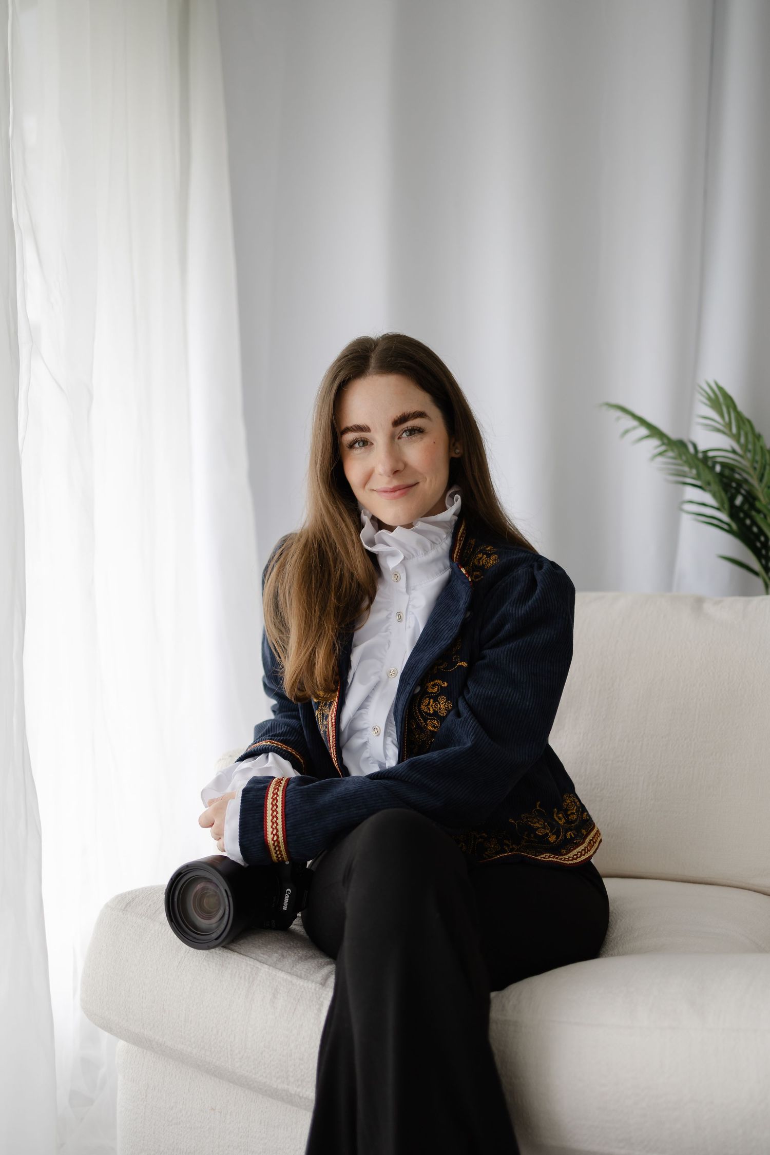 Woman in embroidered navy blazer sits on white sofa holding camera, smiling softly in bright natural light.